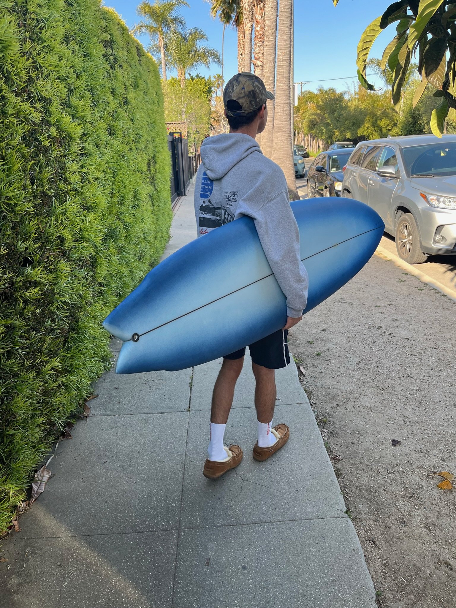 Young man standing on sidewalk holding a blue surfboard, wearing a gray hoodie, camo baseball cap, shorts, white socks, and brown shoes, next to a green hedge and palm trees, with parked cars and a clear blue sky in the background.
