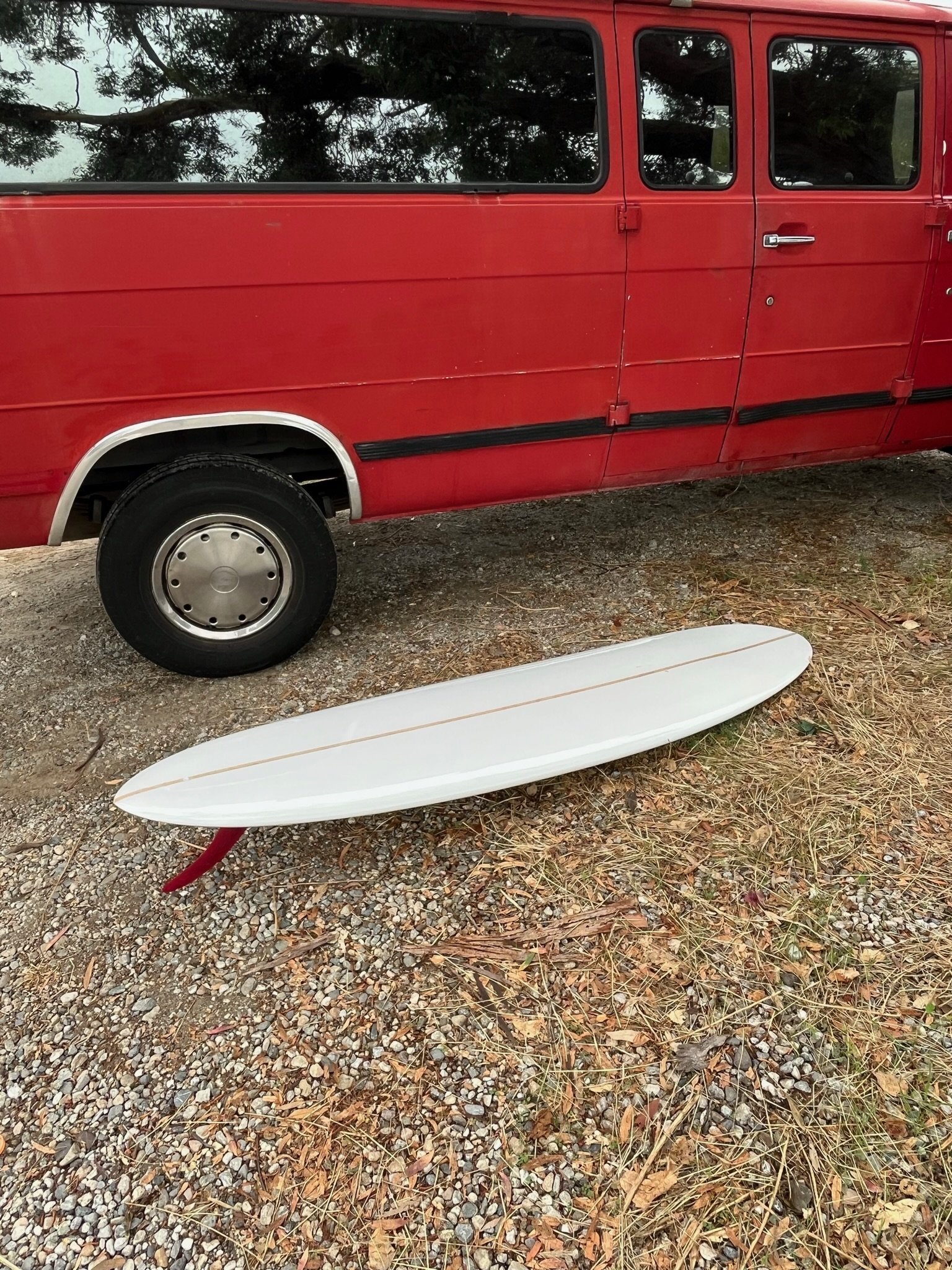 A white surfboard with a red tail lying on the ground amidst small rocks and dried grass, next to a red van parked on a gravel surface during daytime.