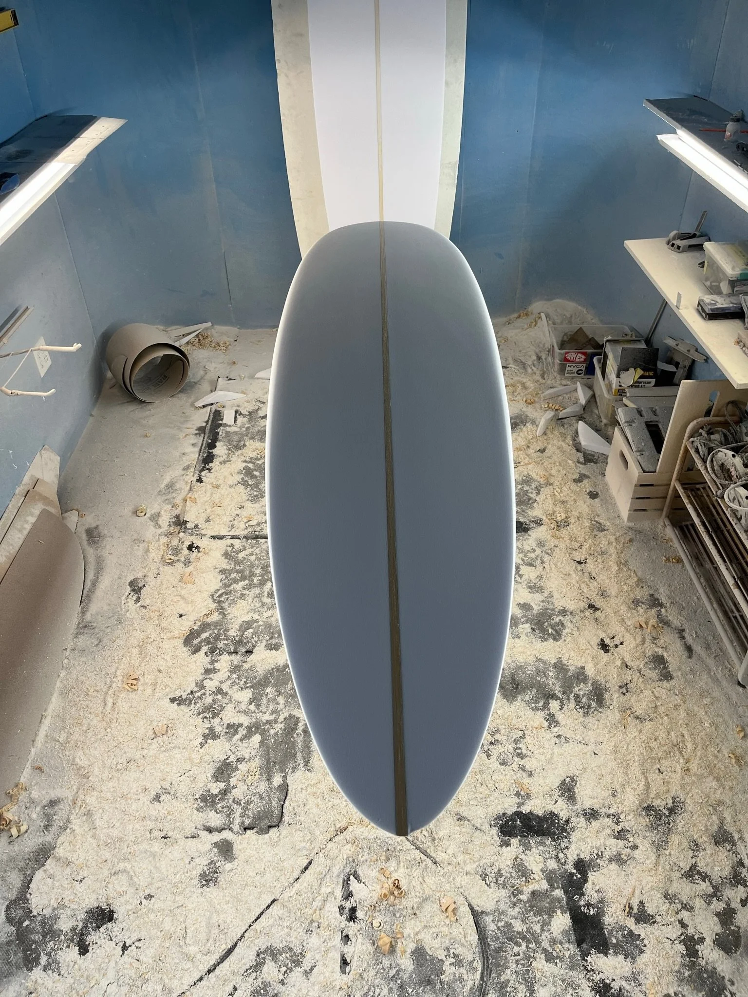 A surfboard standing upright in a room with unfinished, damaged concrete and construction supplies scattered around. The room has blue walls and white shelves with various tools and materials.