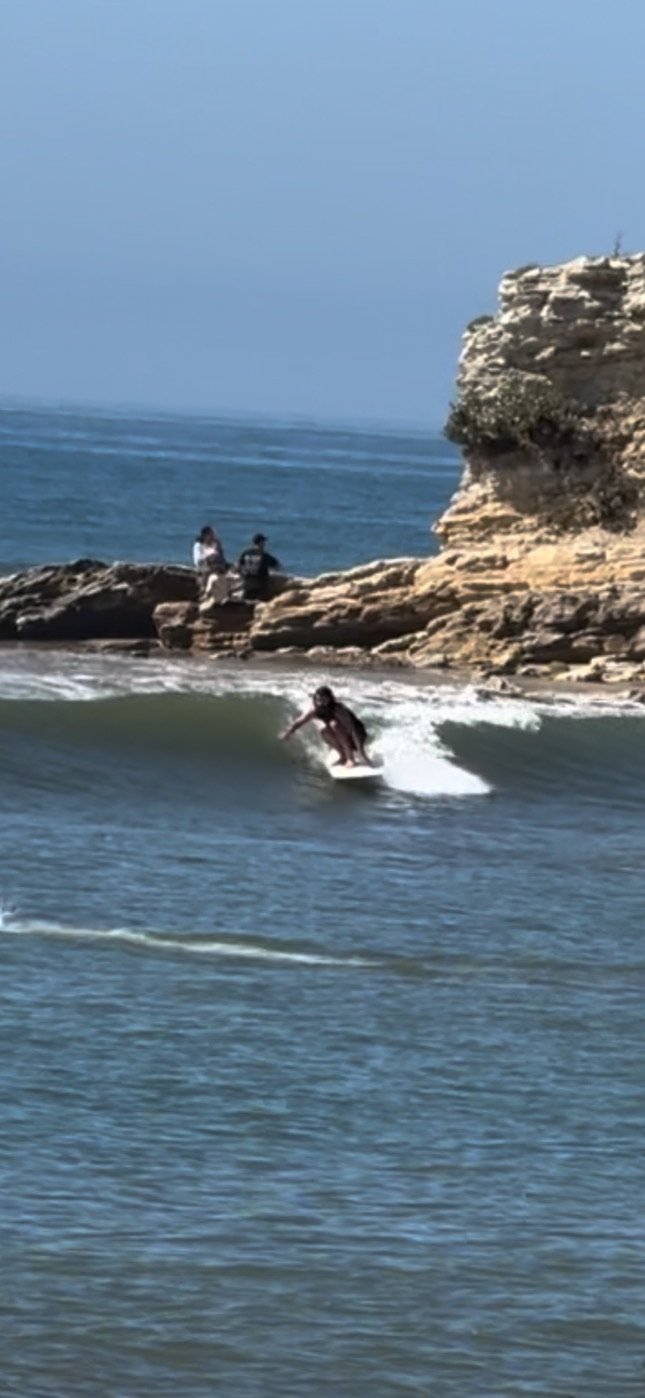 A person surfing on a wave in the ocean near rocky cliffs, with a few spectators sitting on the rocks.