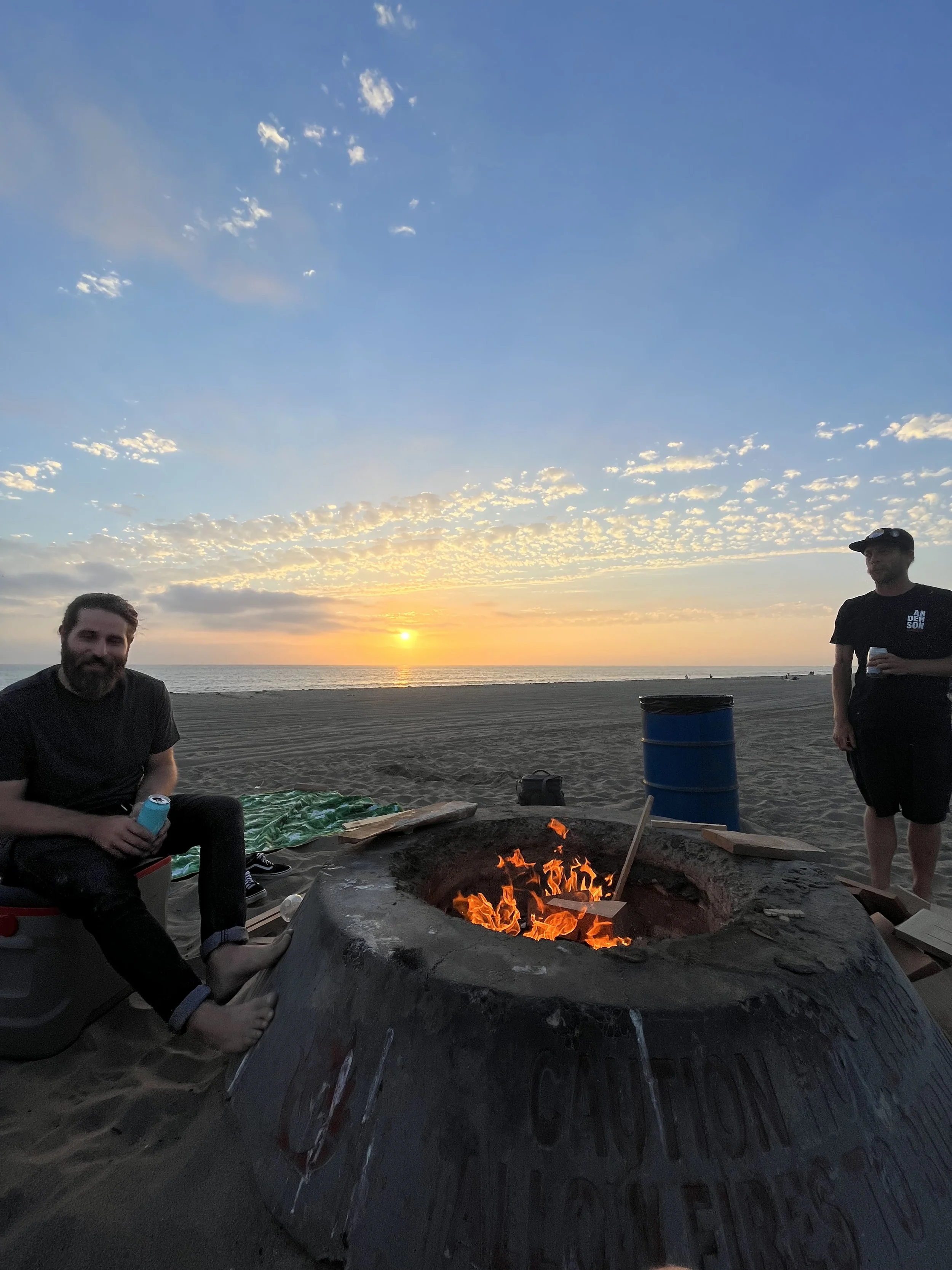 Three men at the beach sitting around a campfire at sunset. One man is sitting on the left holding a drink, another is standing on the right with a drink in hand, and the third man is partially visible on the far right. The sky is partly cloudy with 