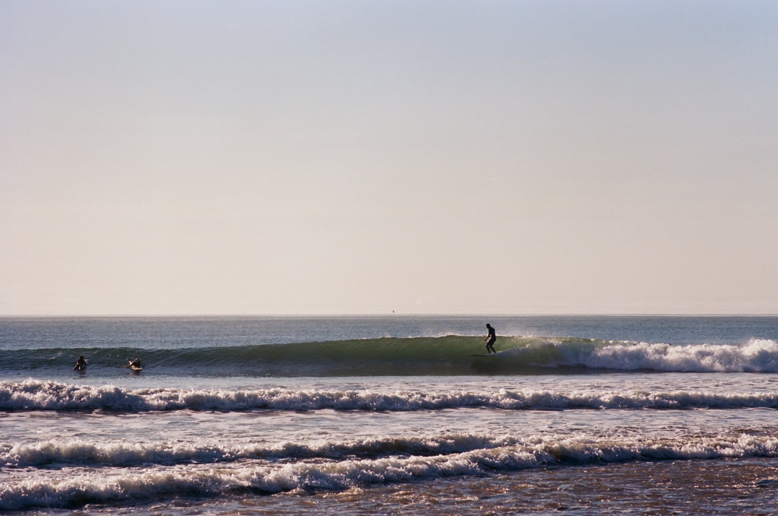 People enjoying the ocean waves, with one person paddleboarding and others swimming or wading in the water, under a clear sky.