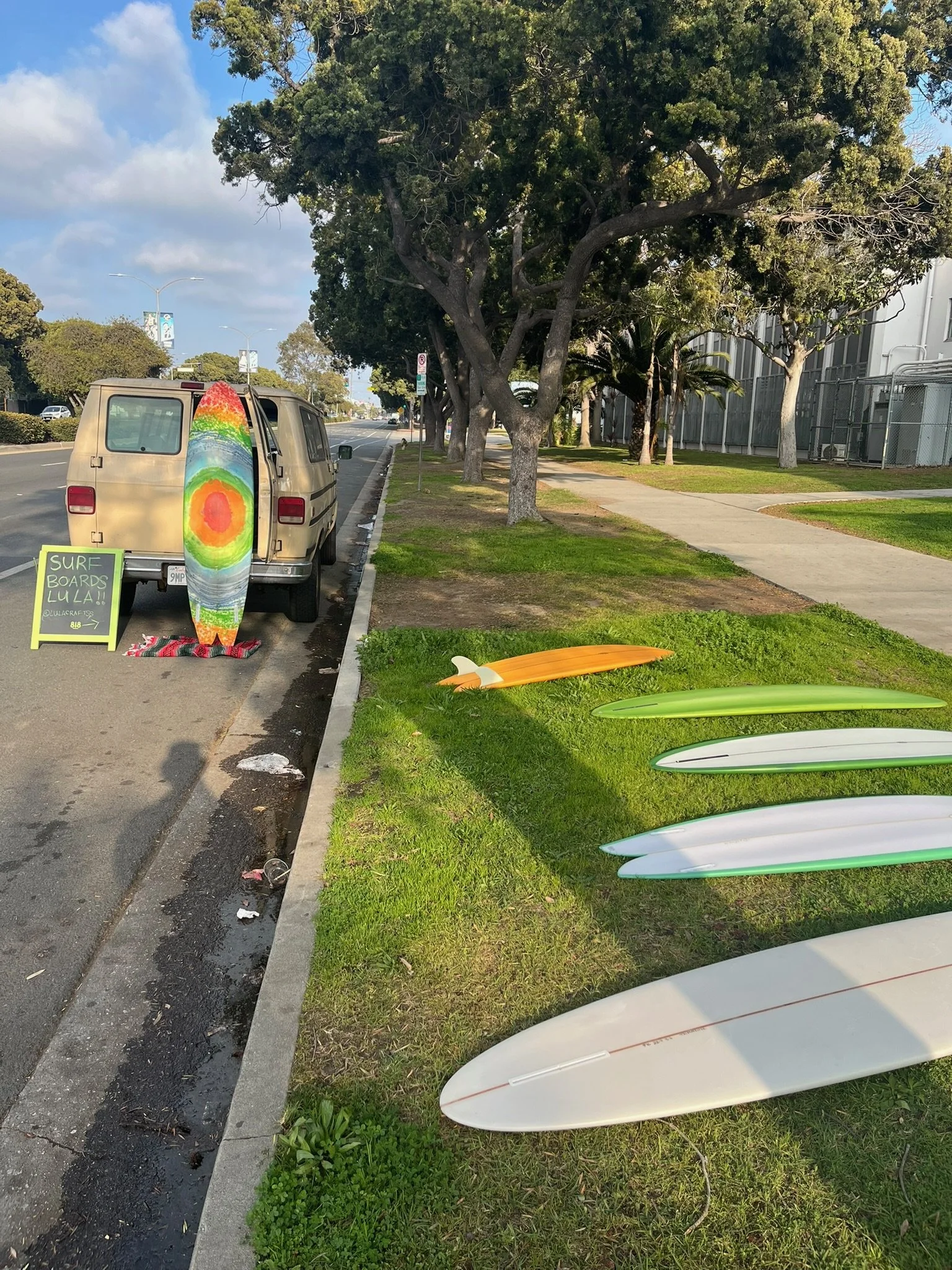 Surfer boards are displayed on the grass next to the sidewalk, with a sign advertising surfboards for sale and a painted surfboard leaning on a vehicle. The vehicle is parked alongside a street with trees, taller modern buildings, and available parki