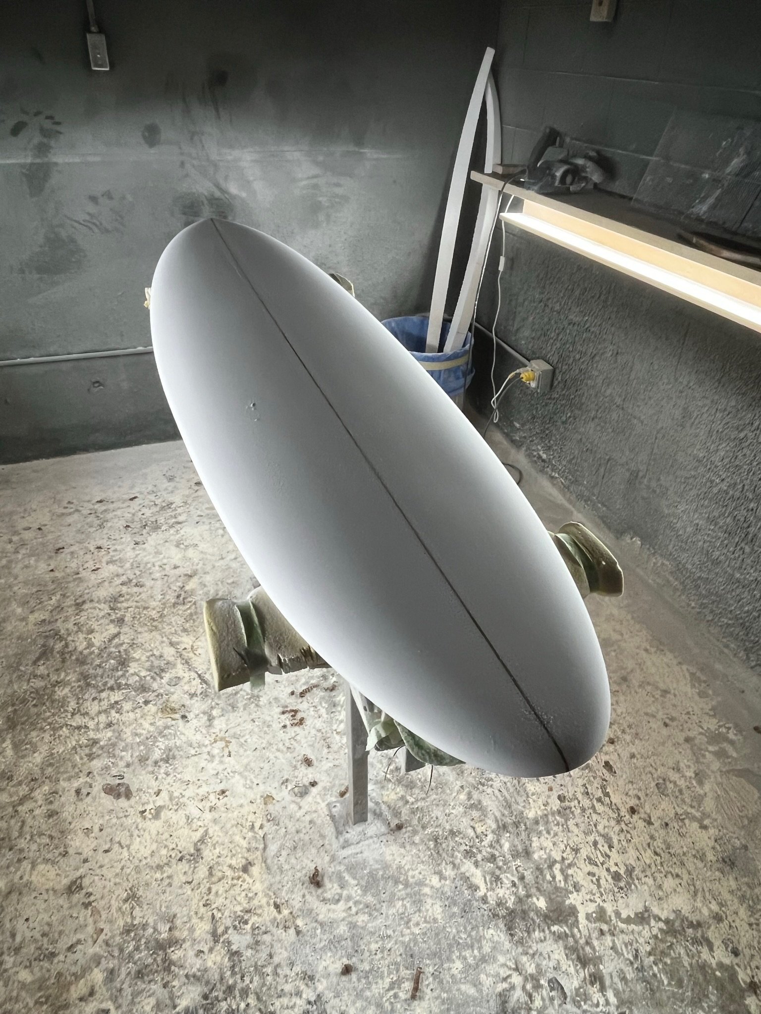 An unfinished surfboard on a work table in a workshop with a concrete floor and a wall with electrical outlets.