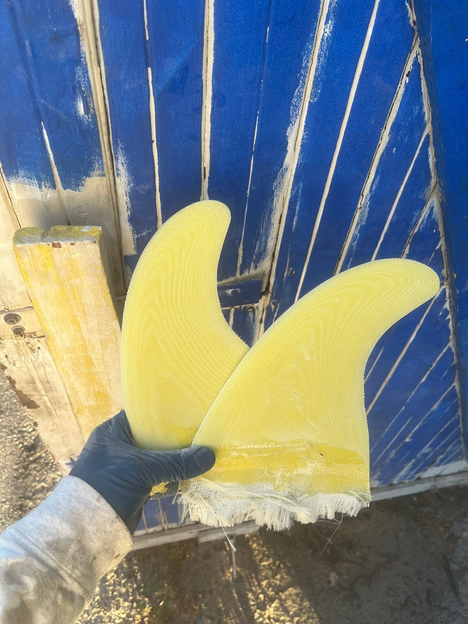 A person wearing a gray sleeve and a black glove holding yellow surfboard fins with a textured surface in front of a distressed blue wooden wall.