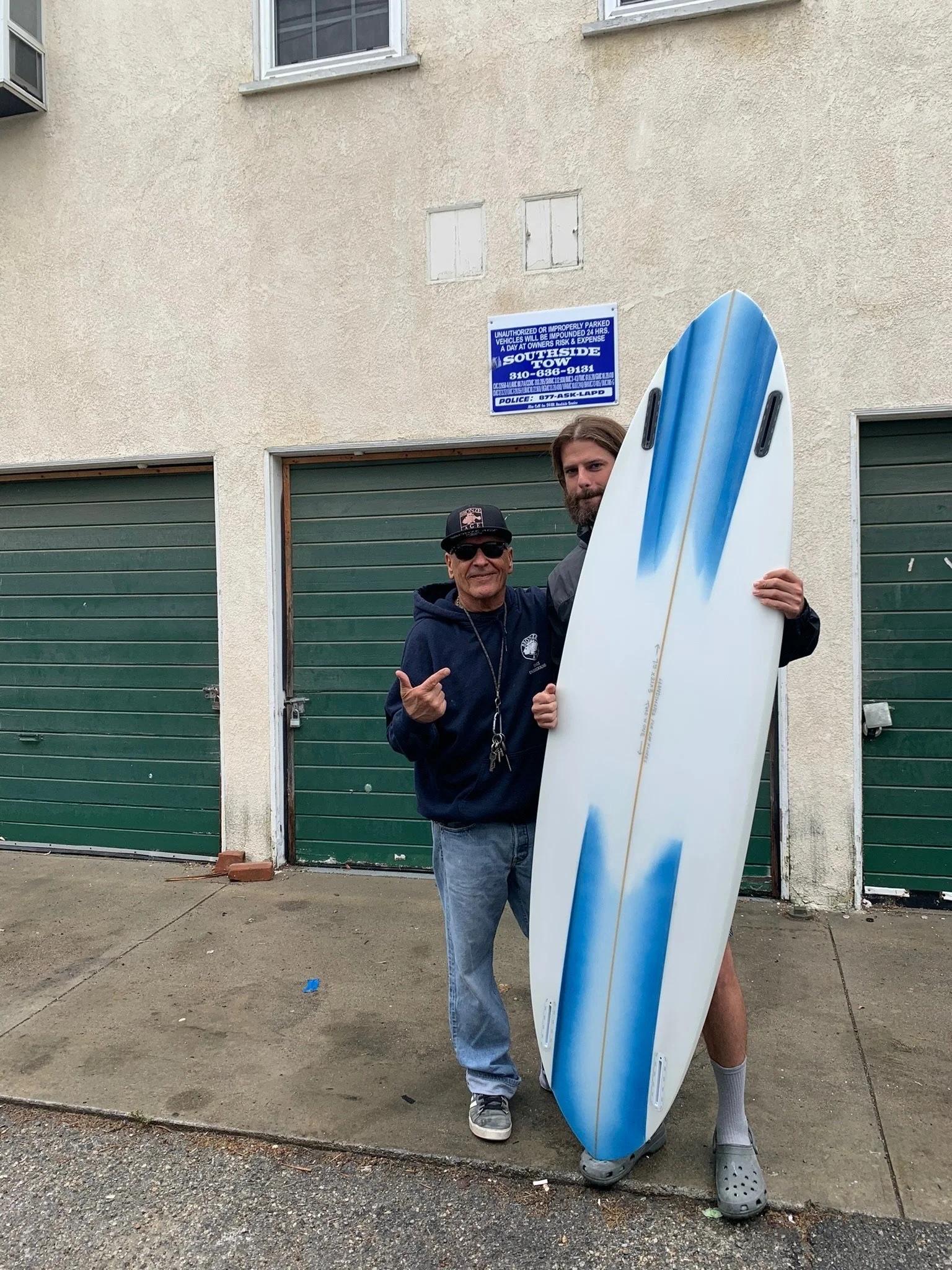 Two men, one older wearing sunglasses and a cap and one younger, holding a white surfboard with blue accents in front of a beige building with green garage doors and a blue parking sign in Venice California.