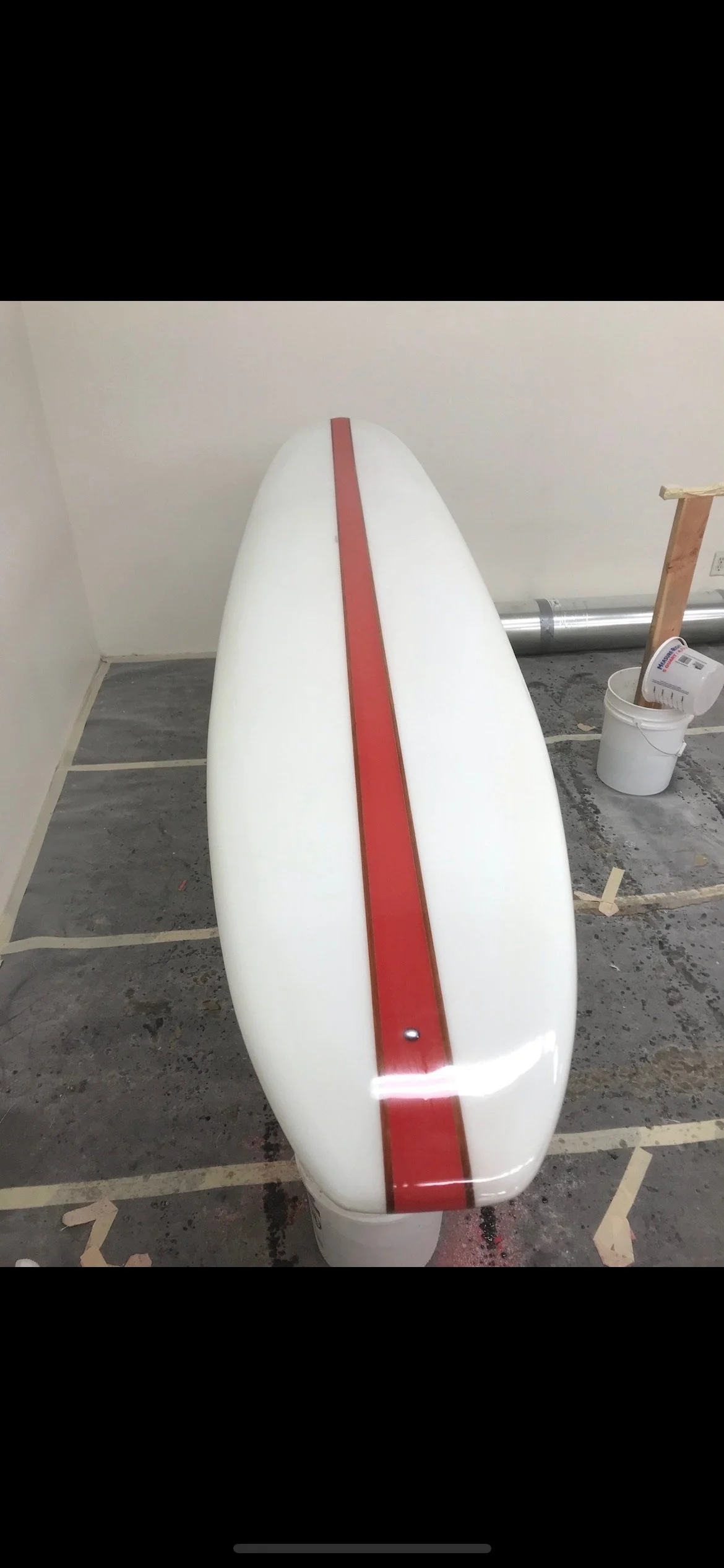 Close-up of a white surfboard with a red stripe down the center, placed on a supporting stand in a workshop.