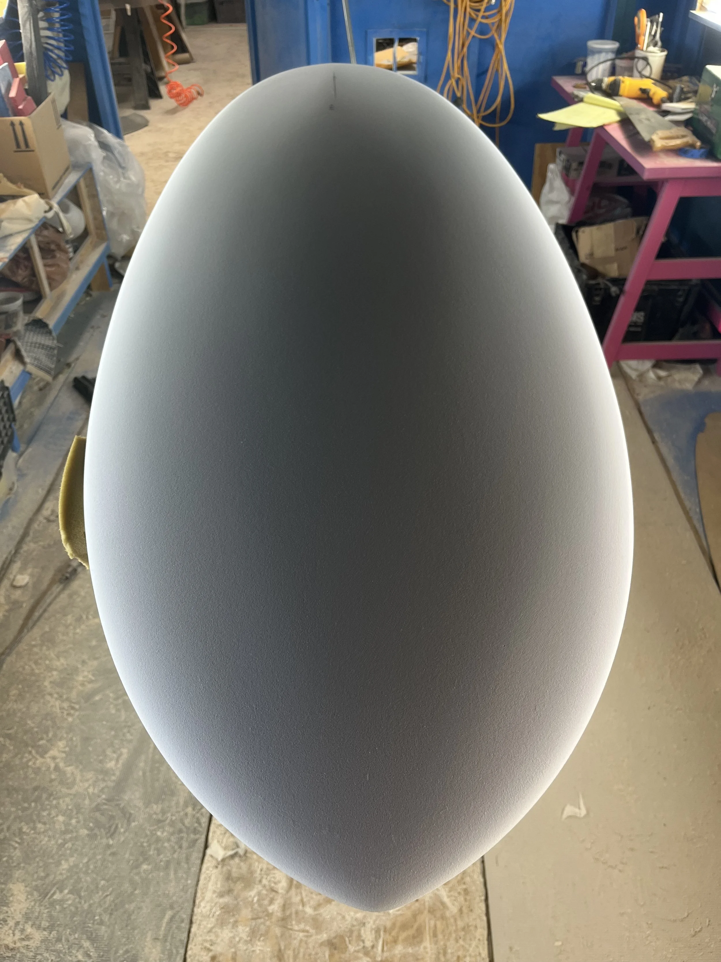Close-up of a smooth, curved, light grey surface, likely a piece of furniture or equipment, in a workshop or tool room with shelves filled with tools and supplies in the background.