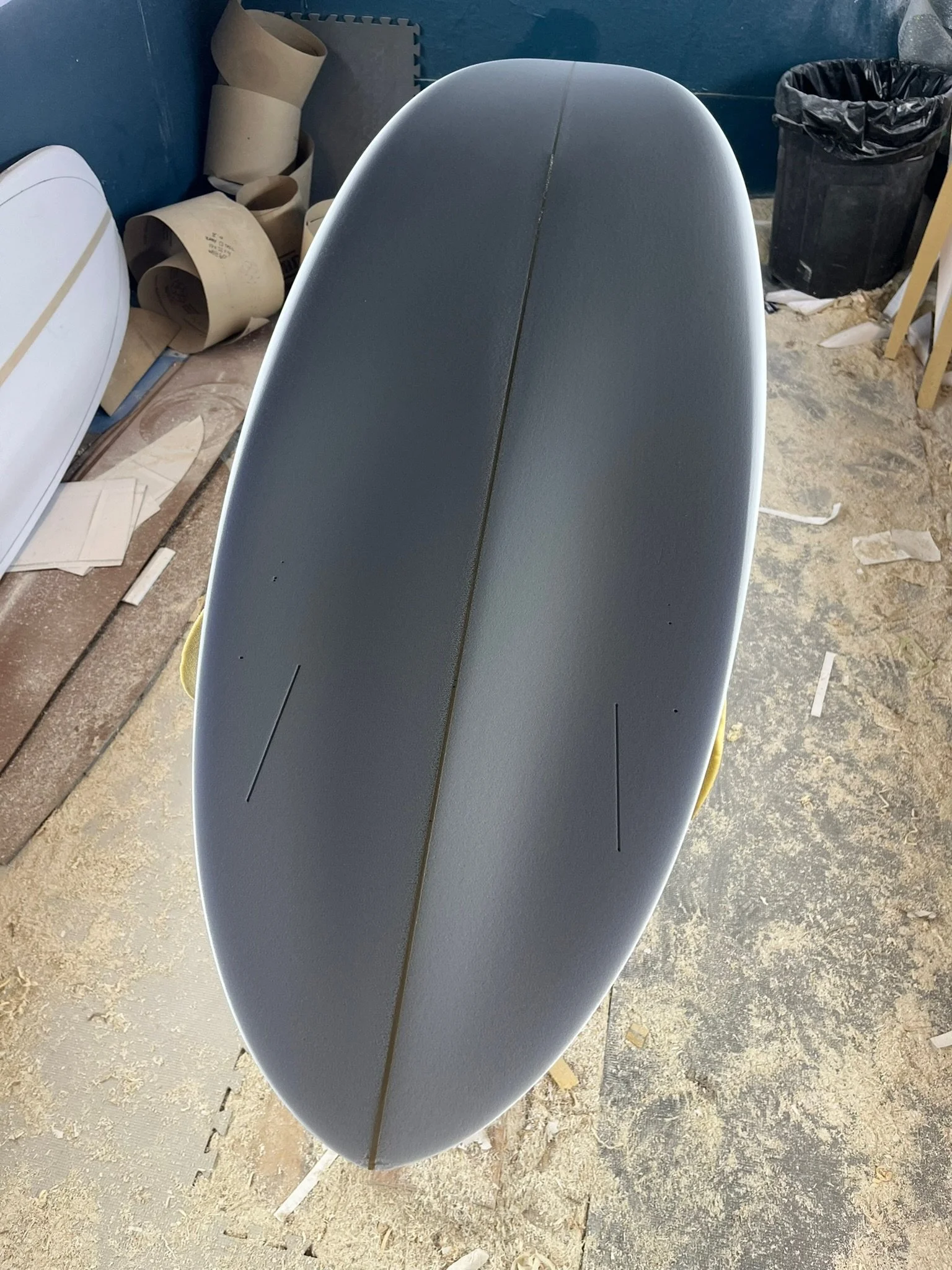 Top view of a surfboard with a gray surface and black line down the center, in a workshop setting with a trash can, cardboard rolls, and foam mats in the background.