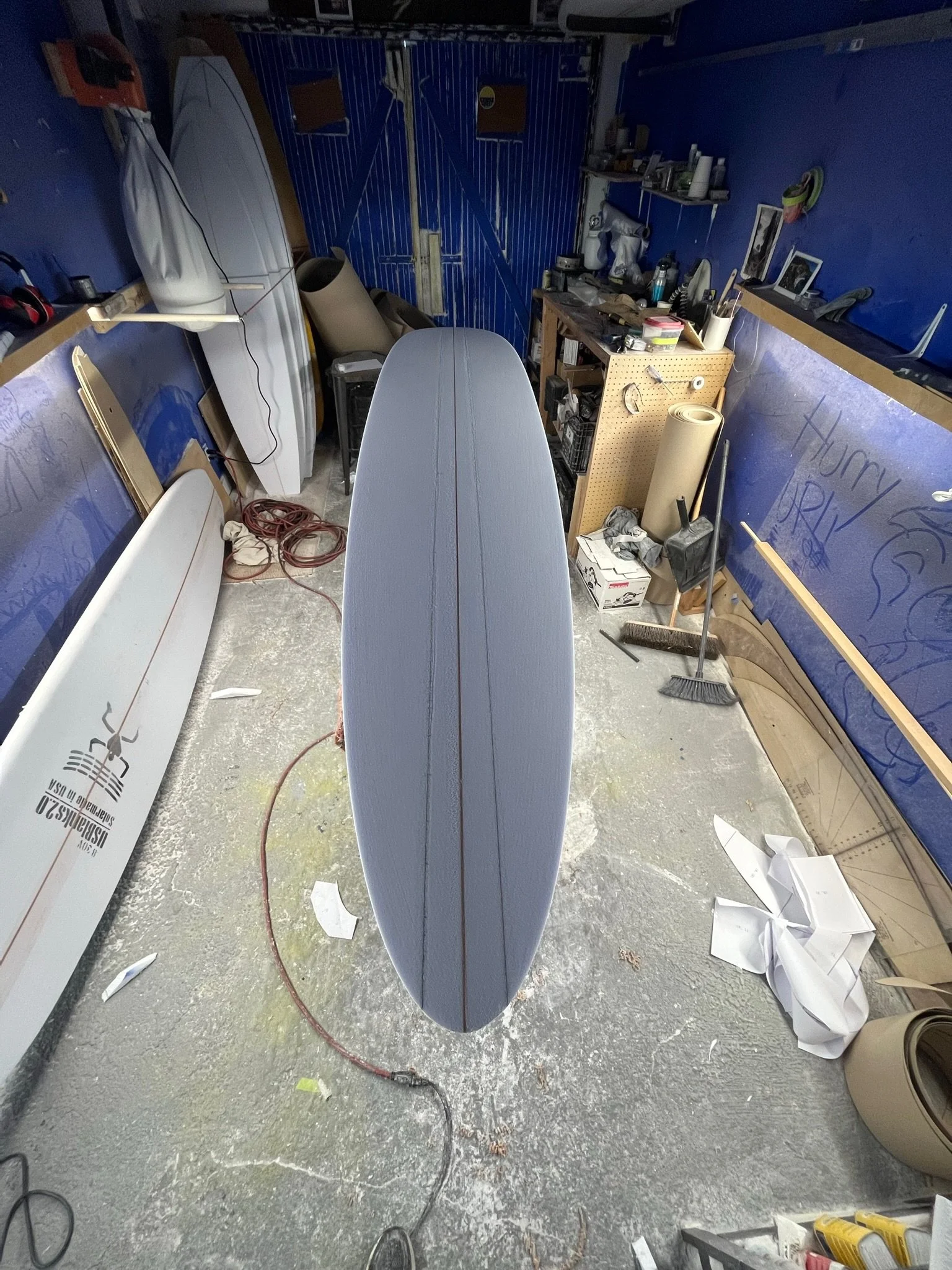 View from above of a surfboard in a workshop or garage, with clutter including tools, wires, and cardboard on the floor and shelves. The surfboard is light gray and positioned vertically in the center of the image.