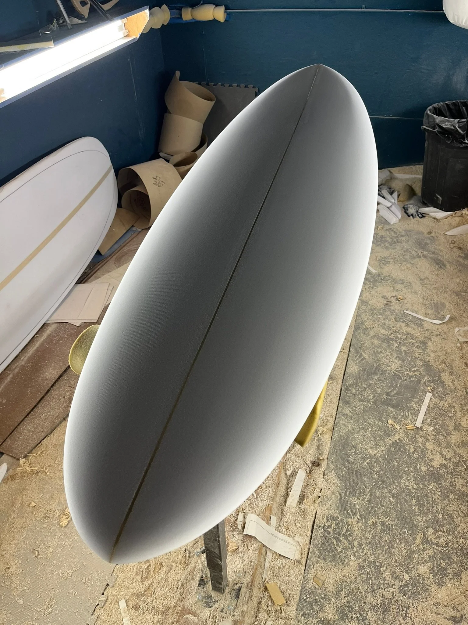 A surfboard leaning on a stand in a workshop with tools, wrapping paper, and a trash bin in the background.