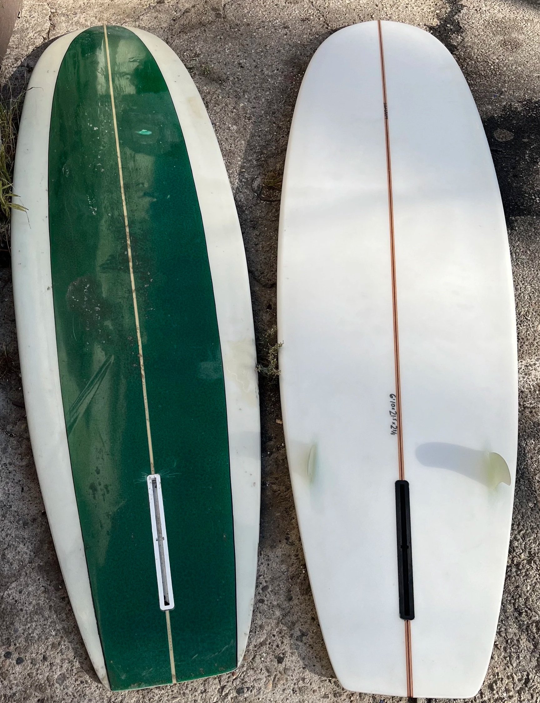 Two surfboards lying on rocky ground, one with a green top and the other plain white with a brown stripe down the center.
