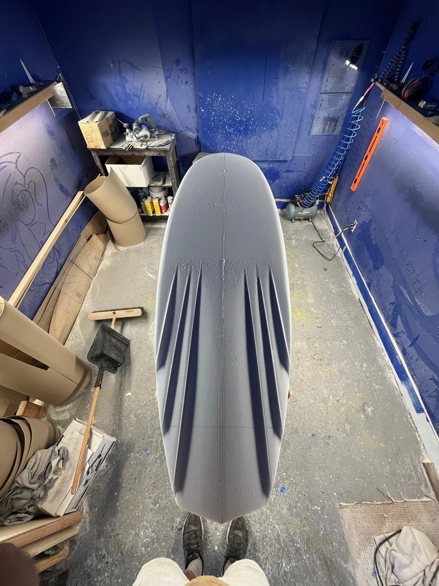 First-person view of a person in a workshop, standing behind a surfboard on a workbench. The workspace has blue walls with spray marks, shelves with tools and supplies, a box, and various equipment on the floor.
