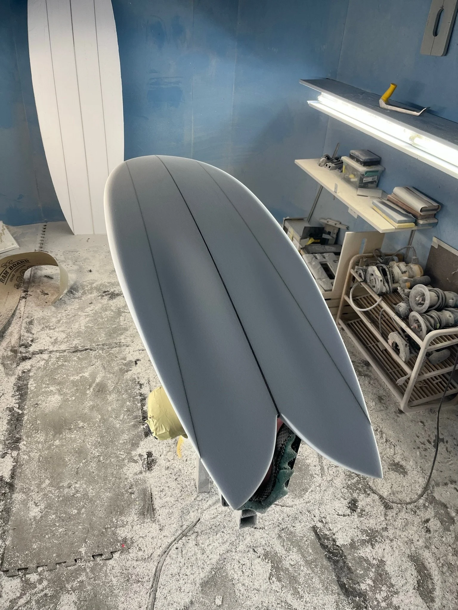 An unfinished surfboard in a workshop with tools, books, and equipment on shelves and tables, and a blue wall in the background.
