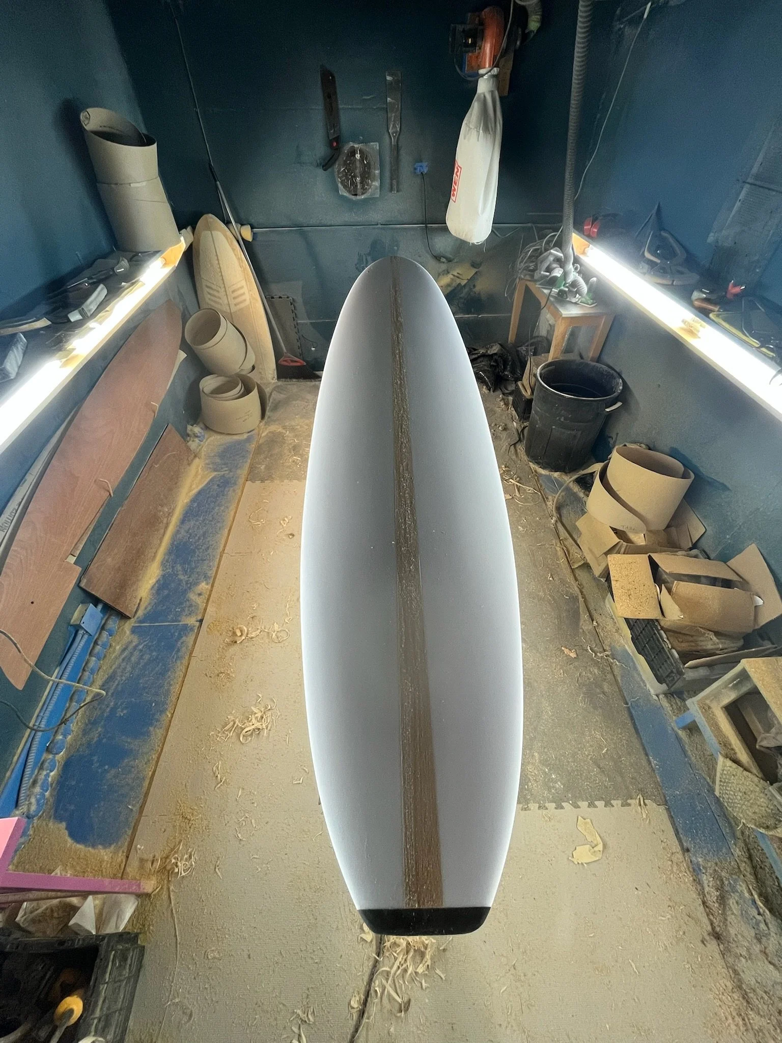 Workshop with a surfboard in the center, tools on the wall, shelves with supplies, and a workbench.