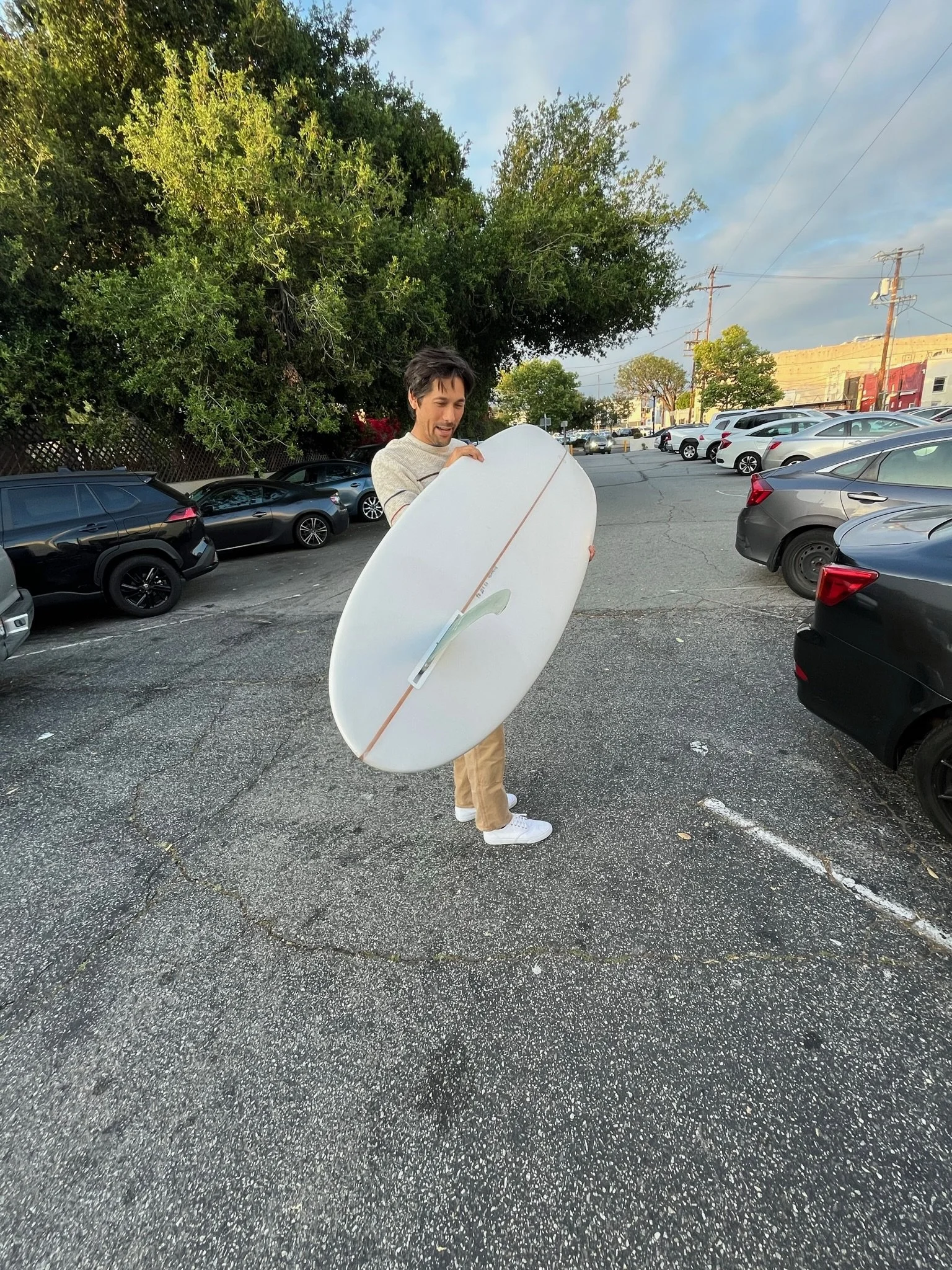 A man standing in a parking lot holding a white surfboard with a plant design, with cars parked on either side and green trees in the background.