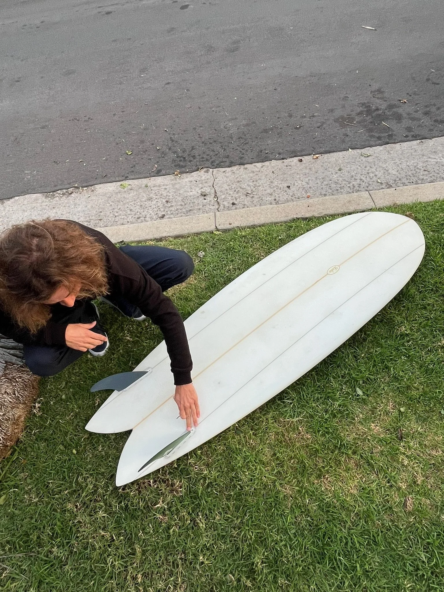 A person crouched on grass near a white surfboard with black fins, pointing at the fins with their right hand. The person has brown hair and is wearing a black shirt and dark pants. The background includes a sidewalk and street.