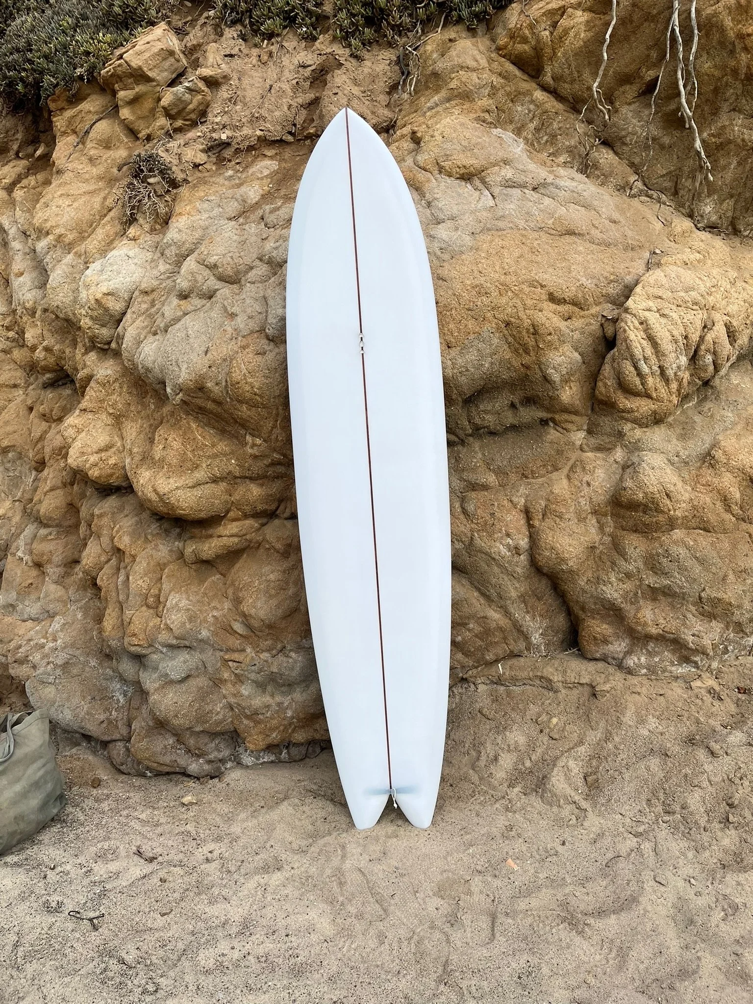 A white surfboard with a red stripe down the middle leaning against a rocky cliff wall at the beach.