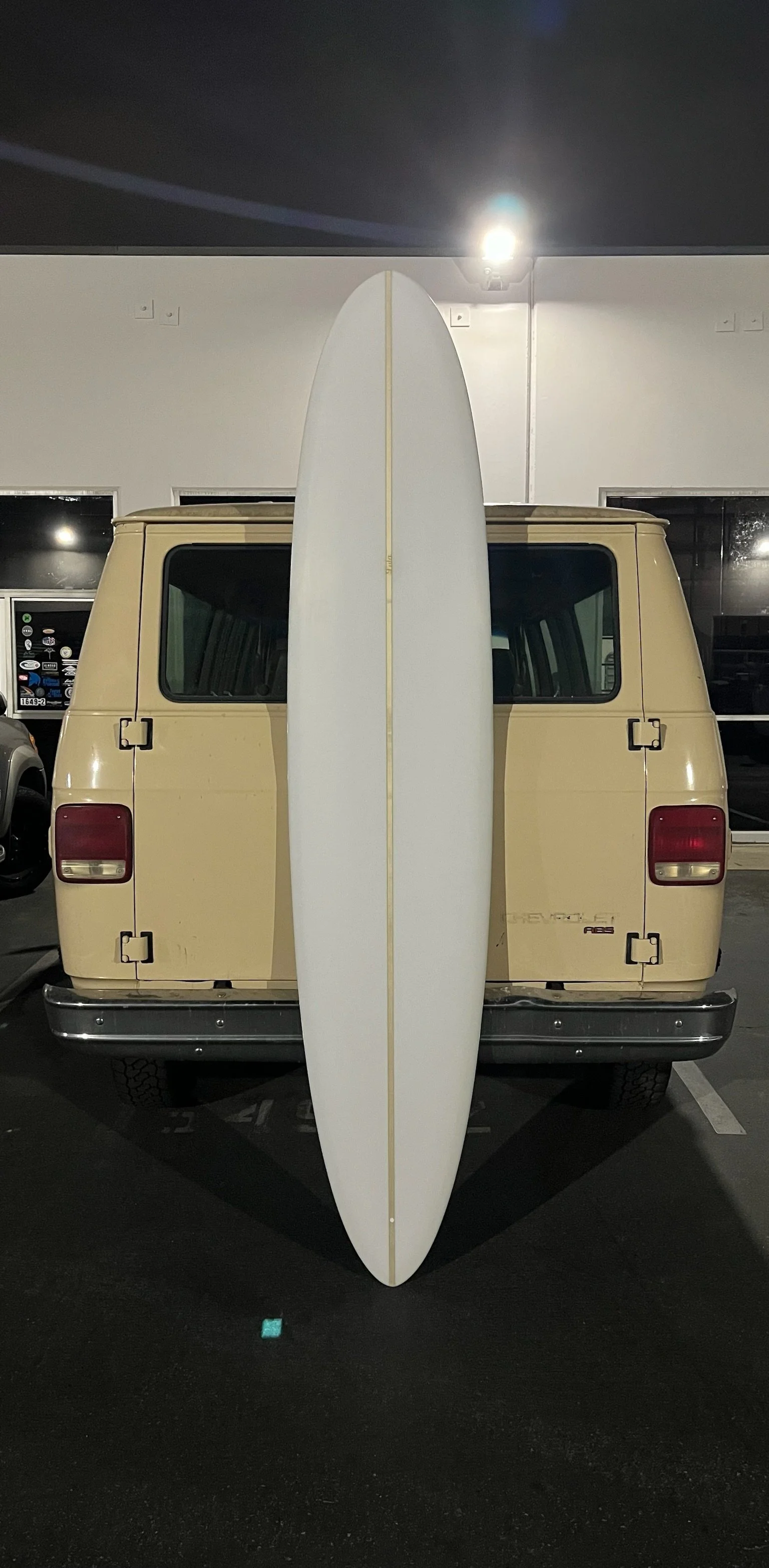 A beige van parked in a parking lot at night, with a white surfboard leaning vertically against the back of the van.