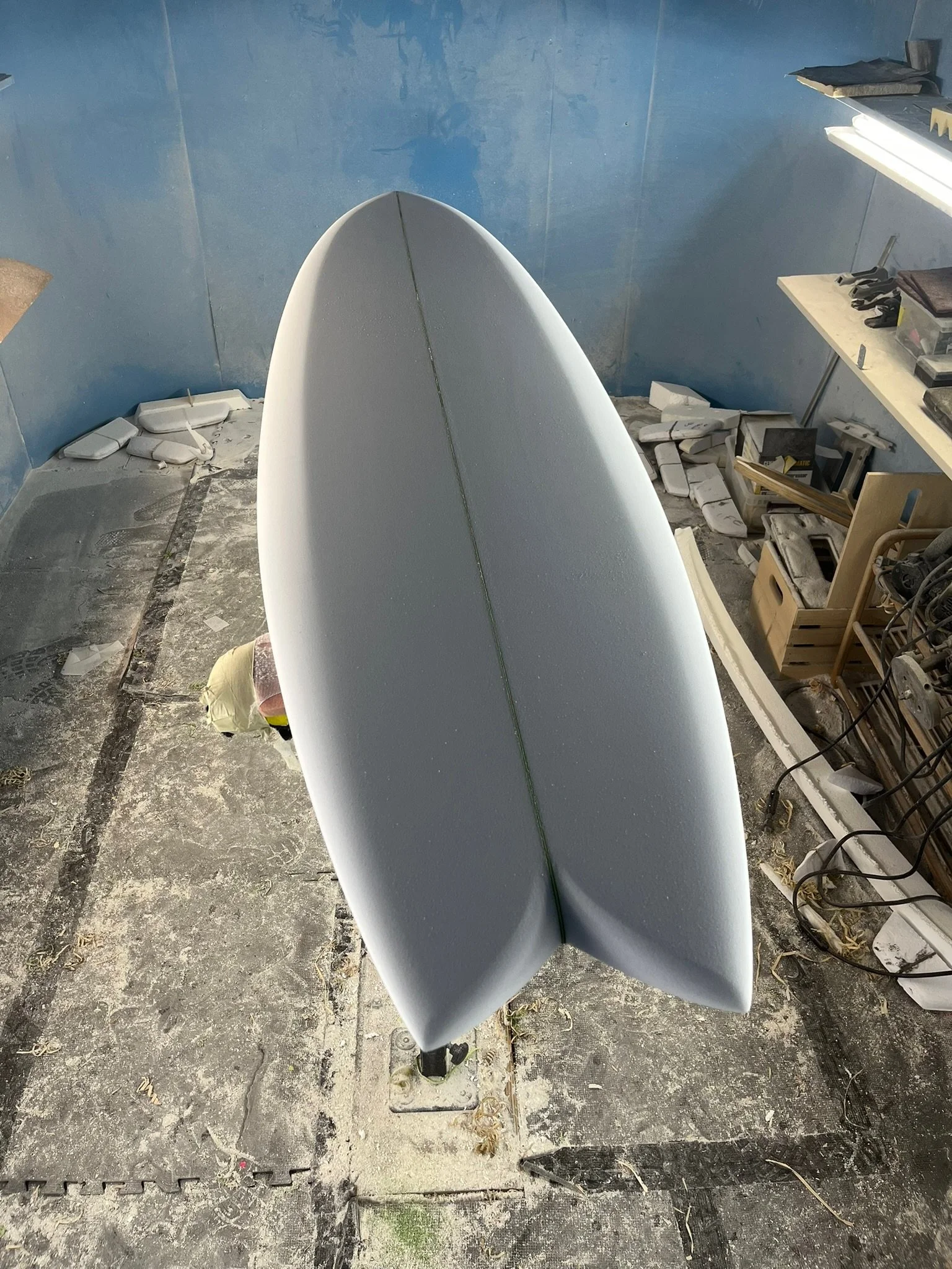A surfboard stands upright on a workbench in a workshop with tools, shelves, and debris on the floor.