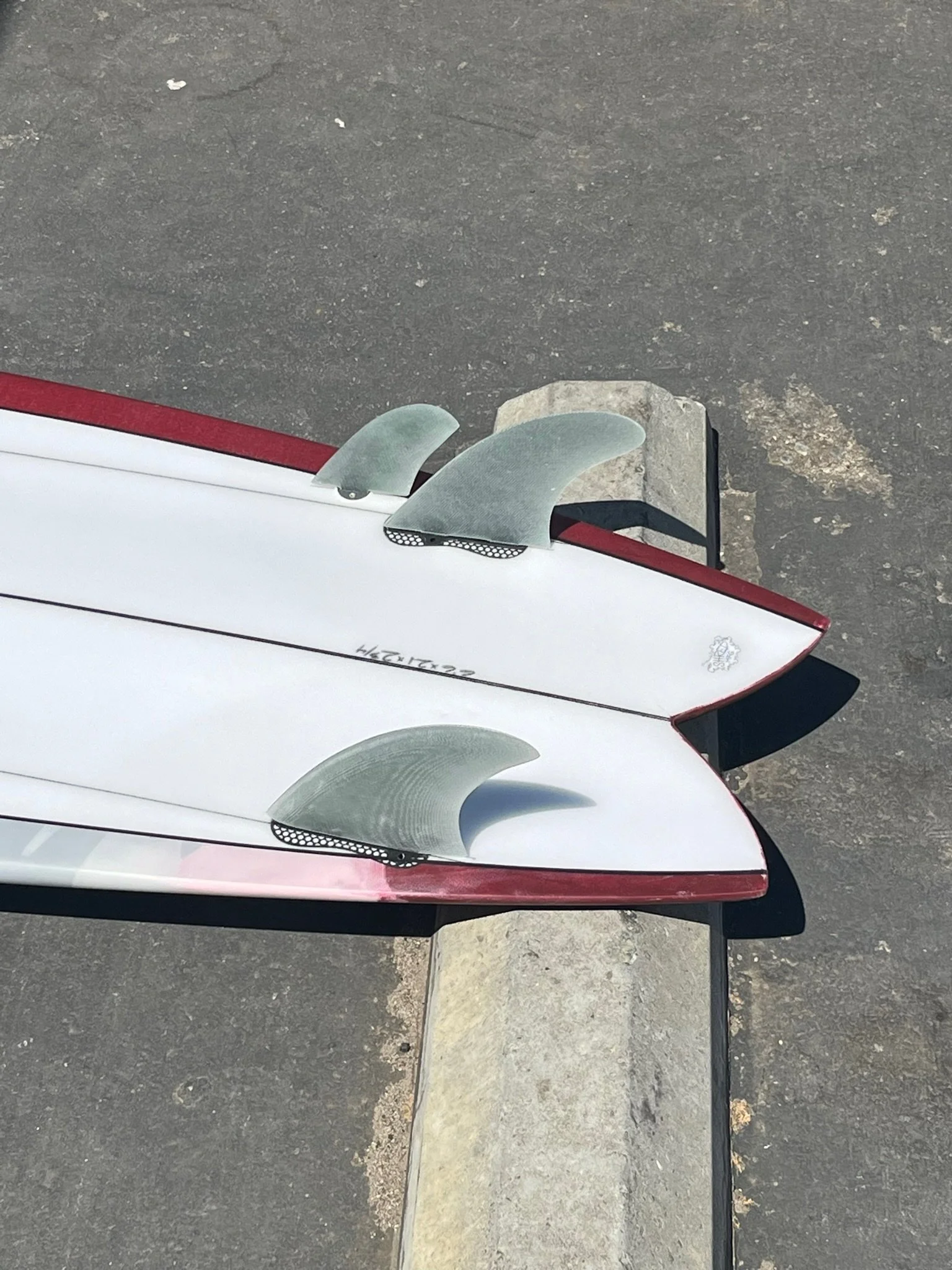 The image shows the front part of two surfboards with fins, one white and one red, placed on a paved ground near a curb.
