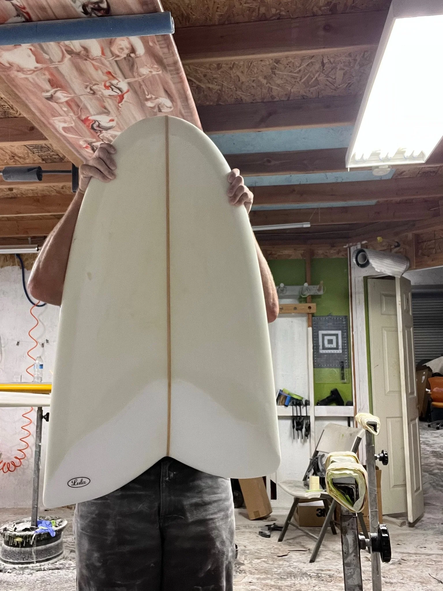 Person holding a white surfboard in a workshop or garage with tools, chairs, and various equipment visible in the background.
