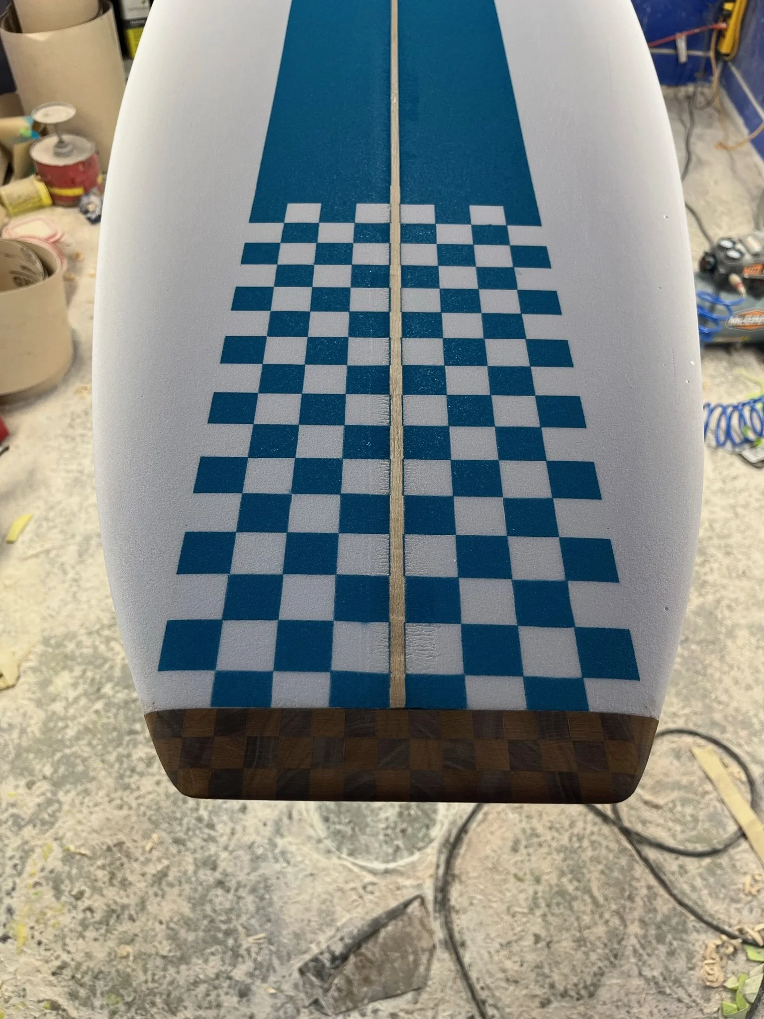 Close-up of a white surfboard with blue checker pattern on top, resting on a wooden stand in a workshop.
