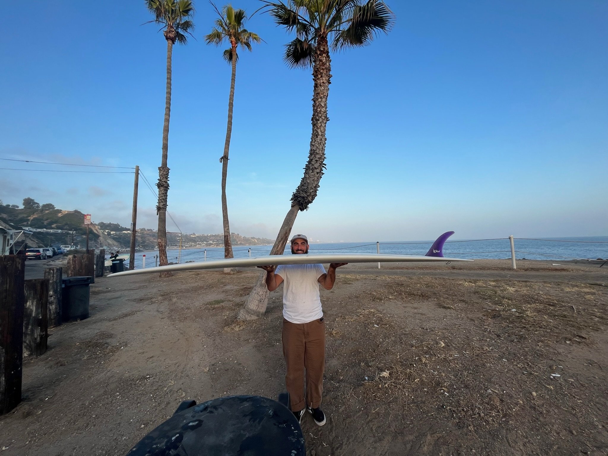 A man standing on a beach holding a surfboard horizontally above his head. There are trees, cars parked along a road, and the ocean in the background.