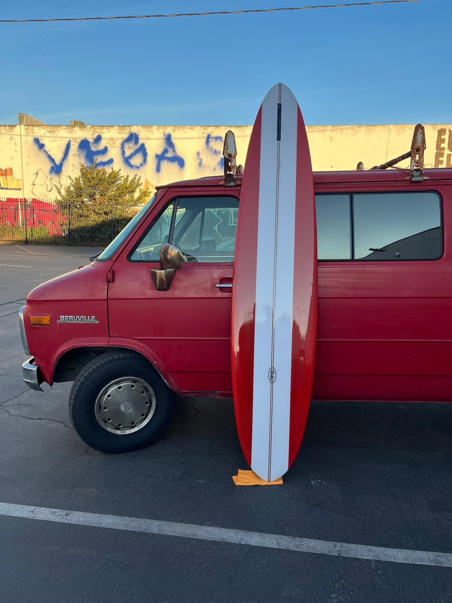 A red van with a surfboard leaning against it parked in a lot. The surfboard is red and white, standing upright with a fin in the middle. A yellow cloth is placed under the bottom of the surfboard. In the background, there is a fence, a tree, and a w