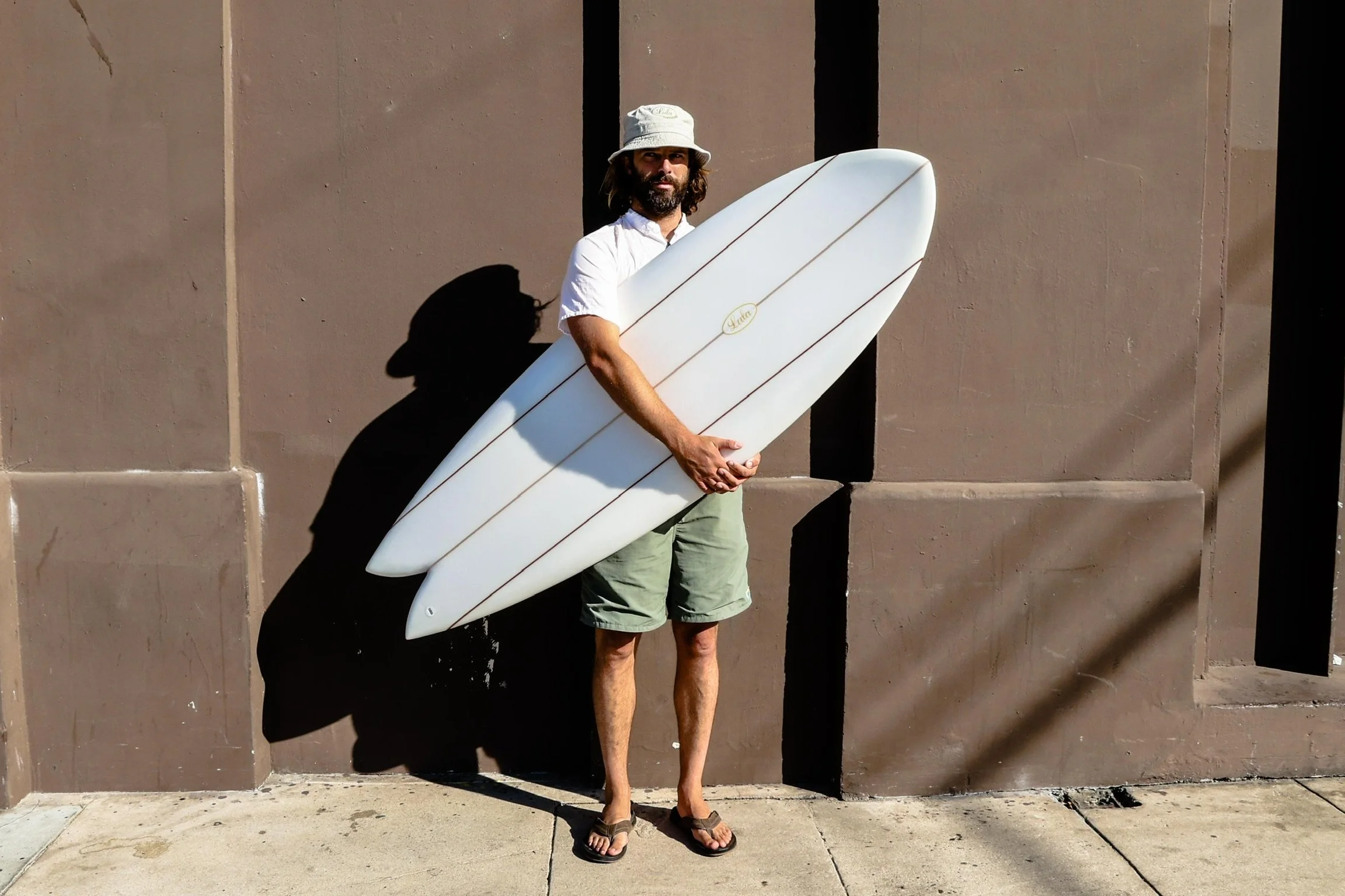Man standing outdoors holding a white surfboard, wearing a bucket hat, white shirt, shorts, and sandals, with a shadow cast on a beige wall behind him.
