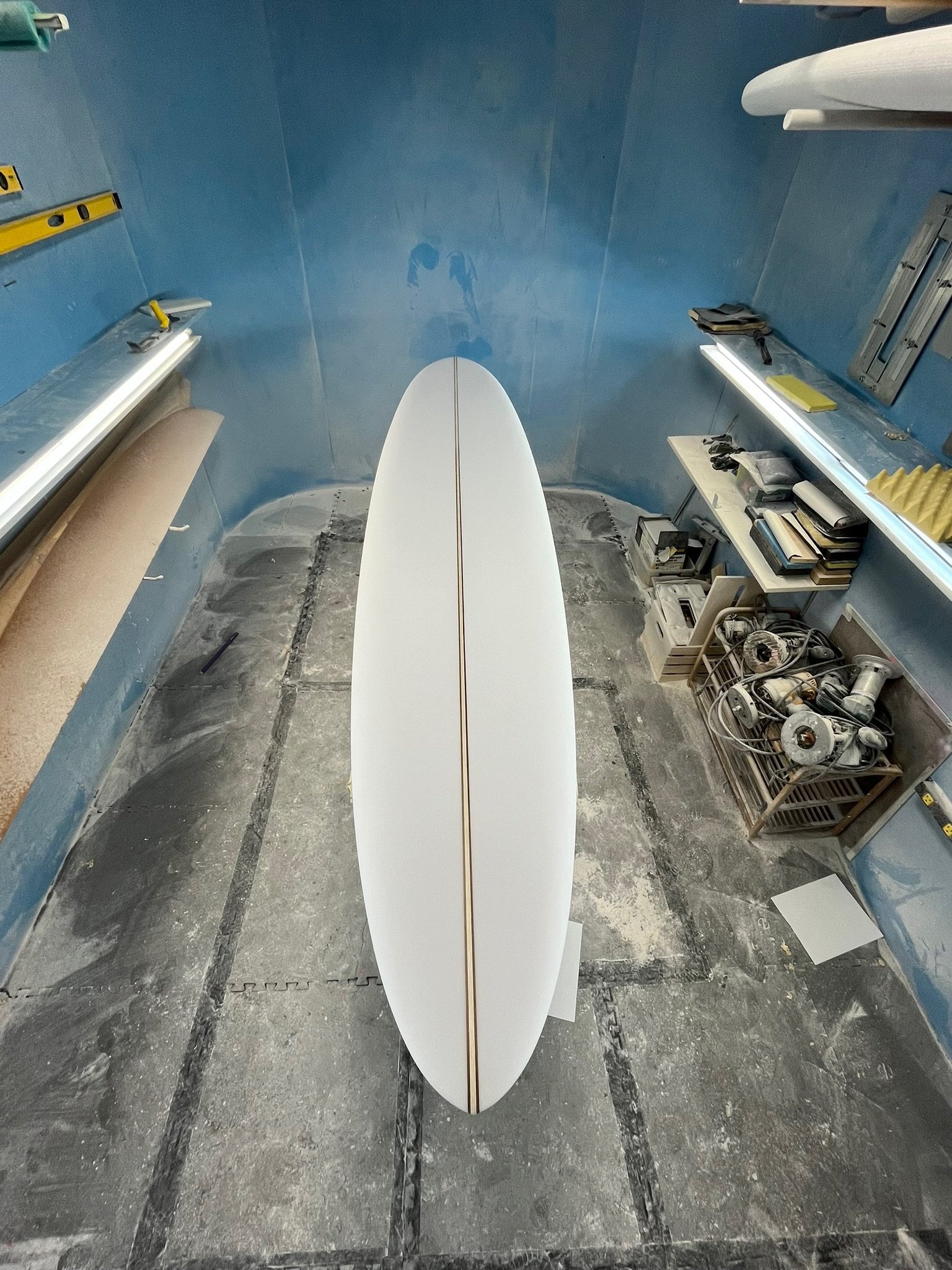 The image shows the front of a white surfboard with a wooden stringer, viewed from the top, standing upright in a room with unfinished concrete flooring and blue walls. Surrounding shelves with books and tools are visible.