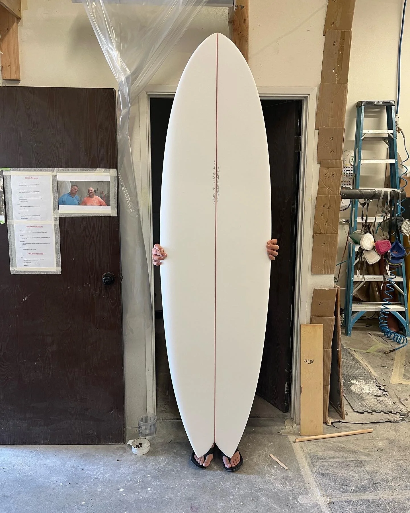 Person holding a white surfboard with a red line down the center, standing in a workshop or garage.