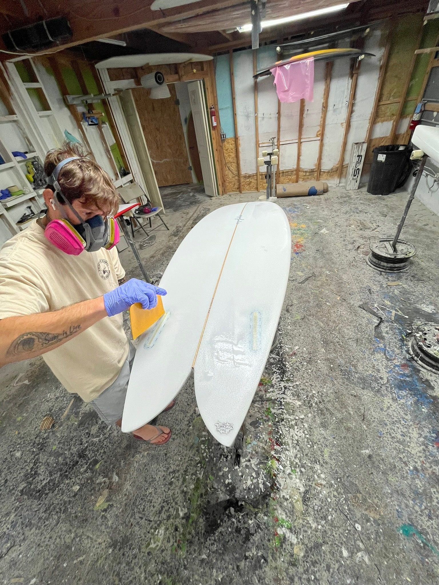 A person wearing a mask, goggles, beige t-shirt, and purple gloves working on shaping a white surfboard in a cluttered garage with unfinished wall panels, surfboards hanging, and paint splatters on the floor.