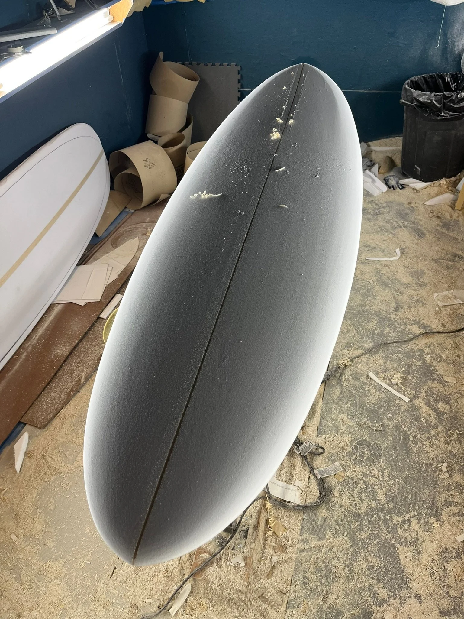 A surfboard with black grip tape on top leaning against a wall in a cluttered garage or workshop. Dust and small debris are on the surfboard's surface. In the background, there are curved foam or cardboard pieces, a trash can, and various scattered i