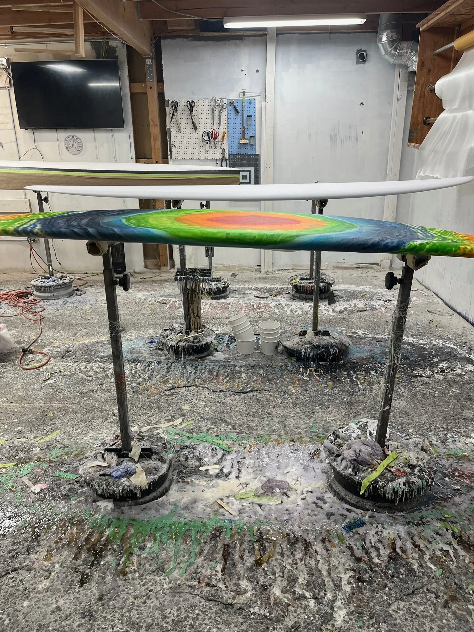 Workshop with surfboards on stands, tools on pegboard, and paint splatters on floor and walls.
