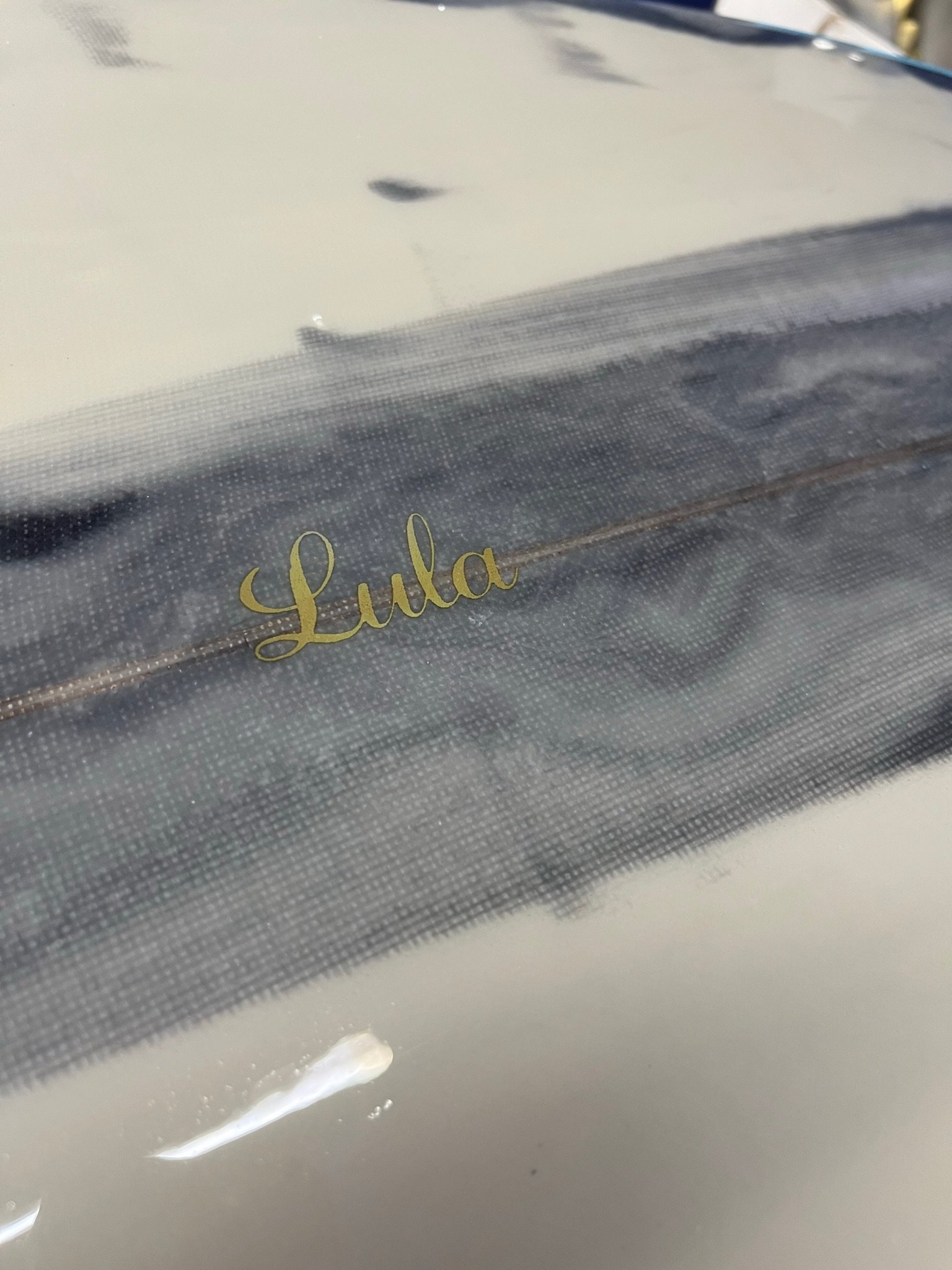 The image shows glossy surfboard deck with gold text that reads "Lula,".