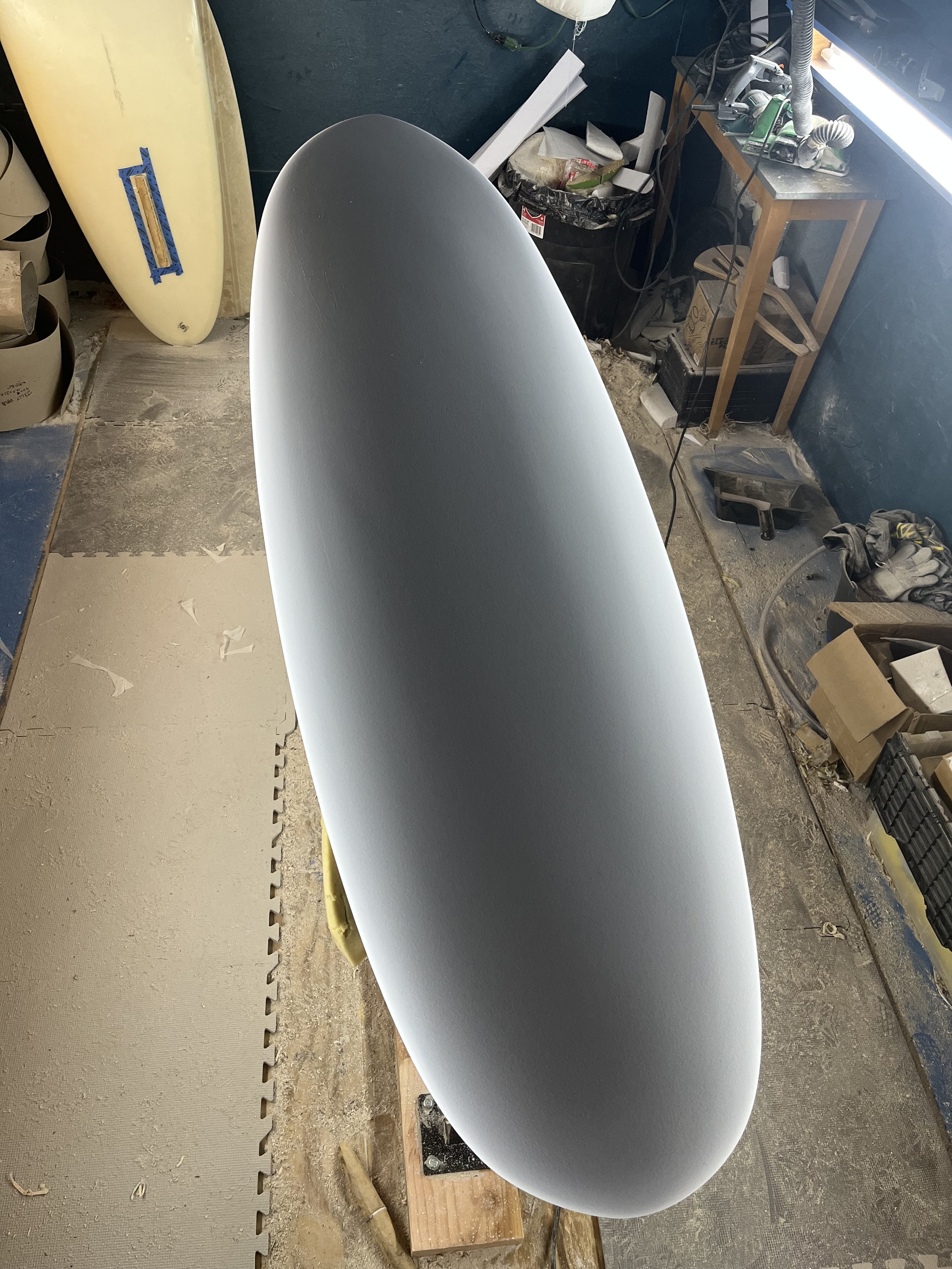 A surfboard in a workshop surrounded by tools, boxes, and workshop materials.