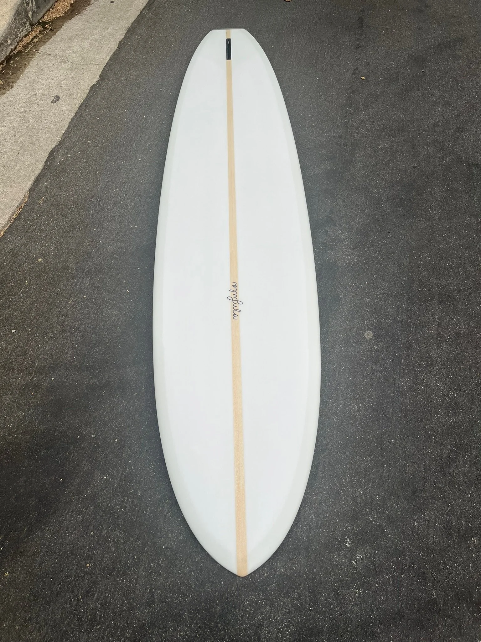 A white surfboard with a tan stripe running down the center, lying on a paved street surface next to a concrete curb, with a small signature near the bottom.