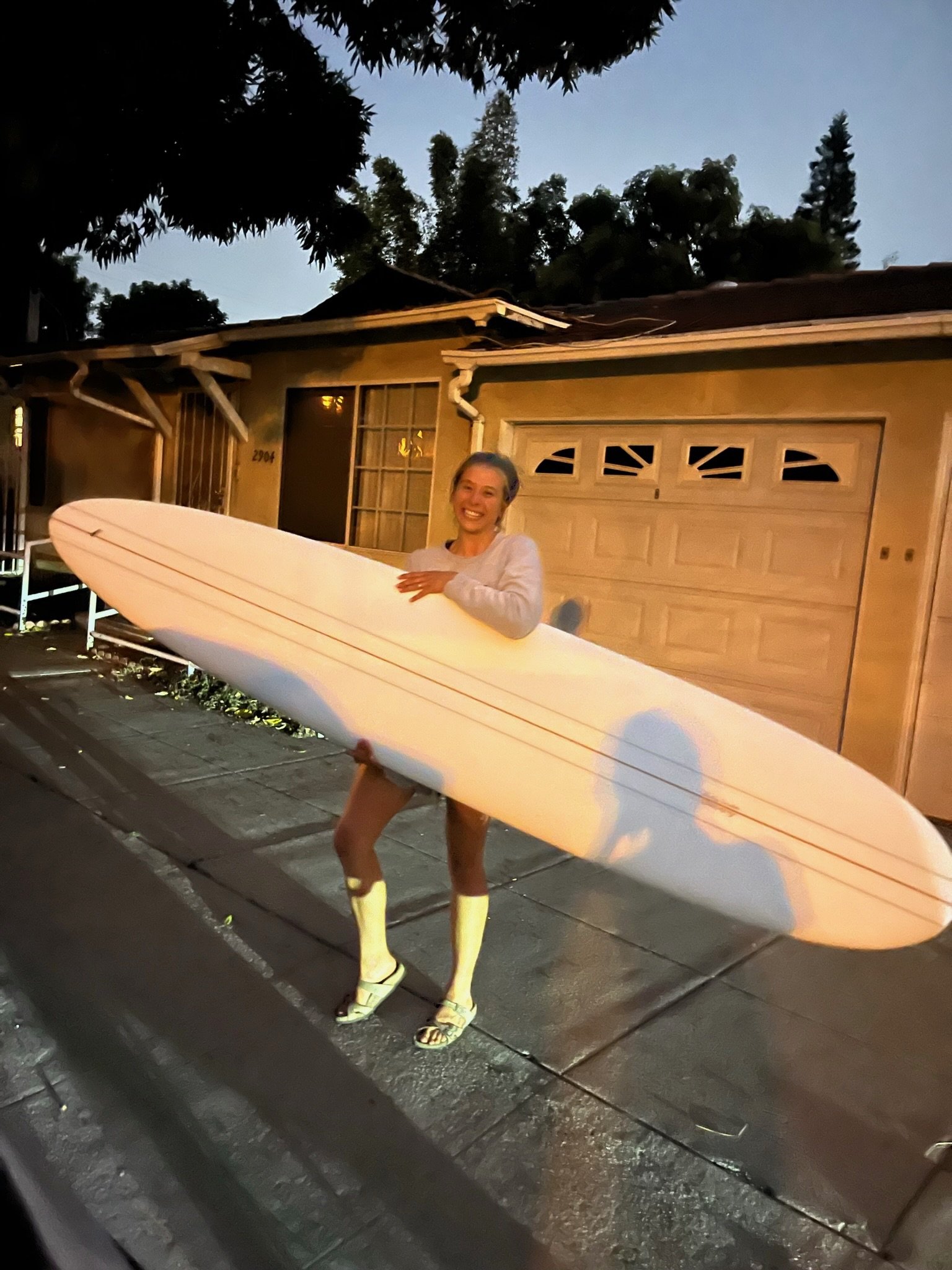 Person holding a surfboard, standing outside of a house at night, smiling, wearing a white jacket and yellow boots.