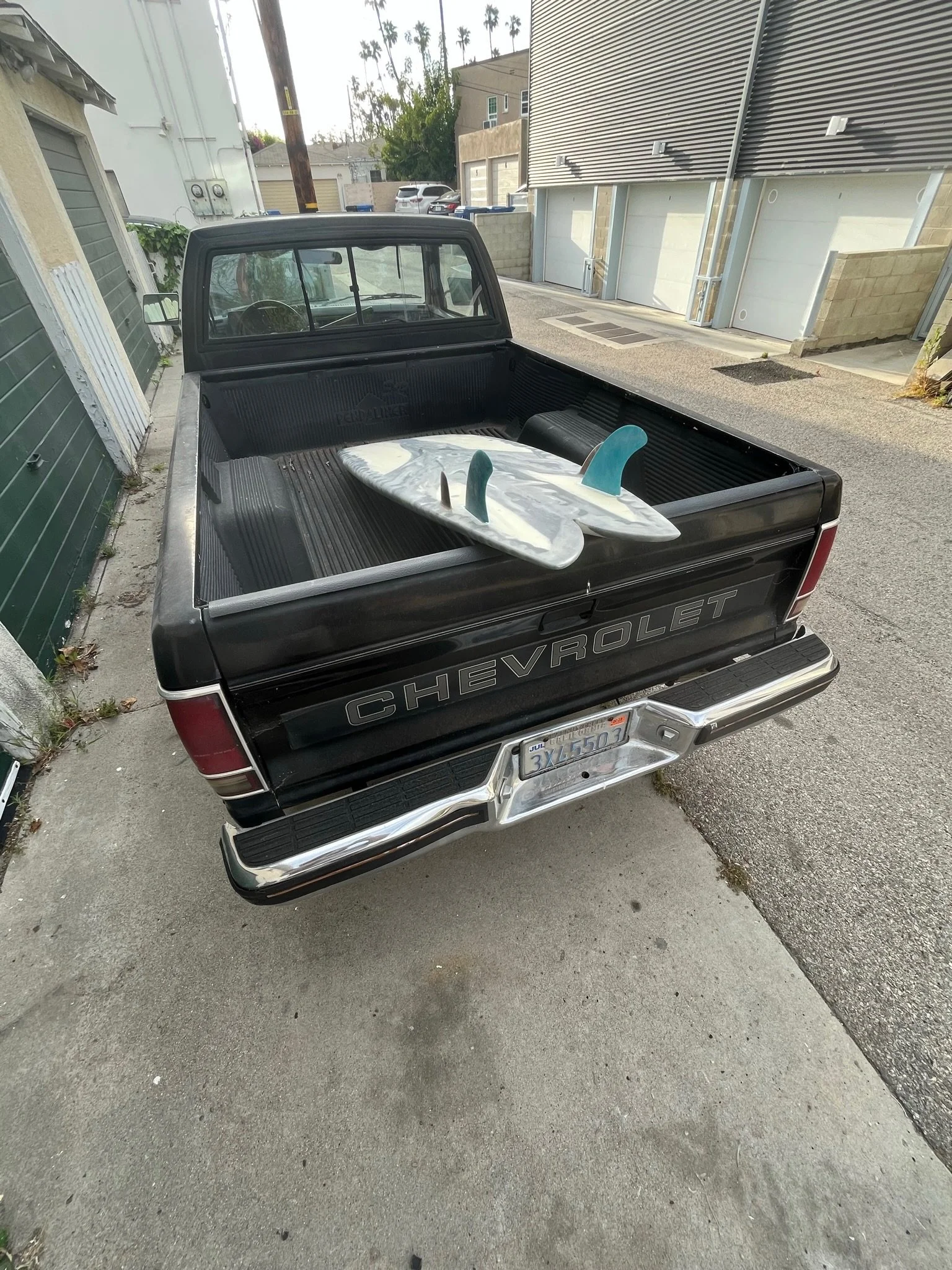 Black Chevrolet pickup truck with two surfboards in the bed, parked on a sidewalk in front of a building, with a green garage door to the left and residential buildings in the background.