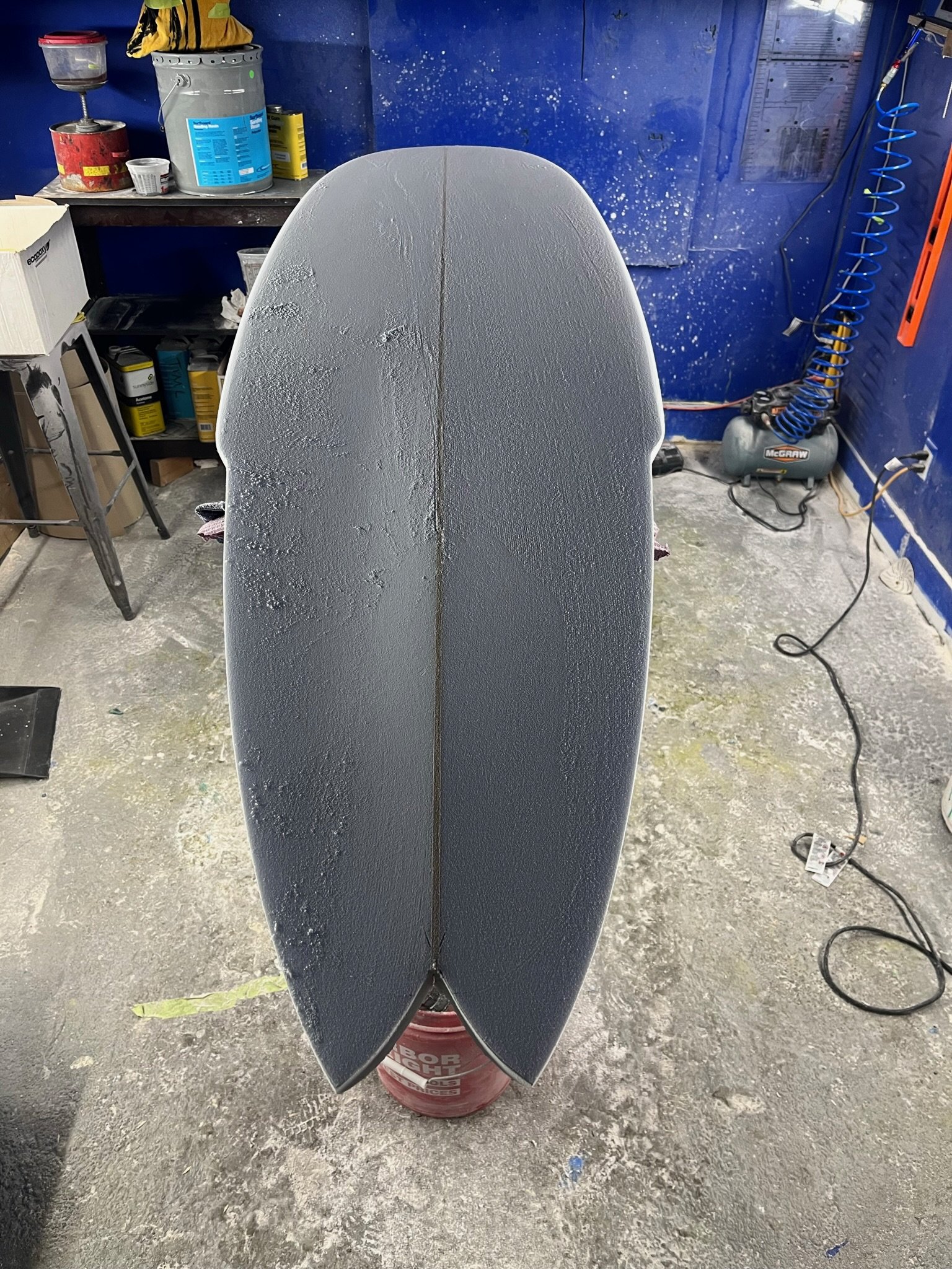 A skateboard deck with a textured grip surface leaning against a red bucket in a workshop with tools and supplies in the background.