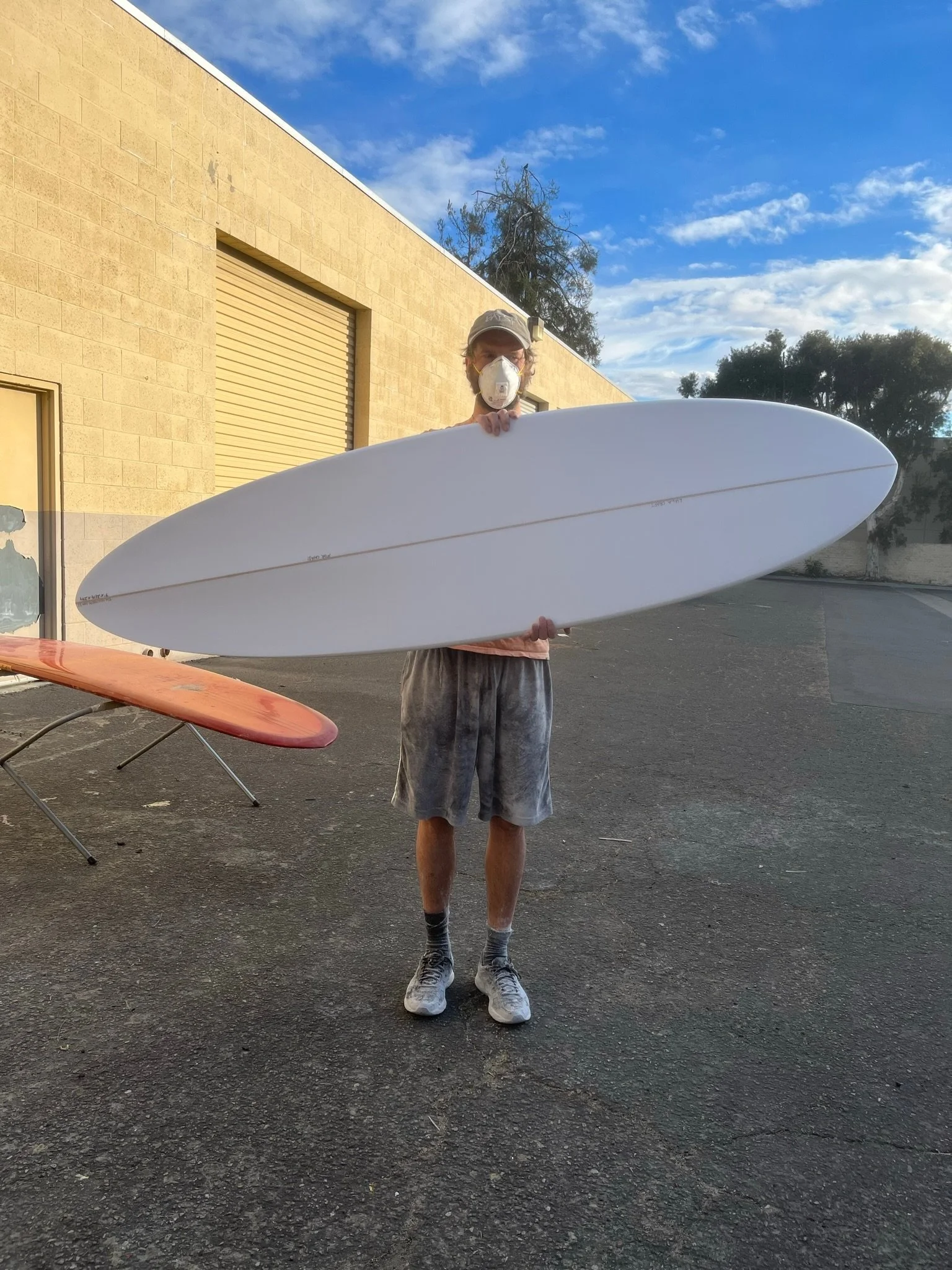 A person wearing a cap, face mask, T-shirt, shorts, and sneakers, holding a white surfboard outdoors in front of a beige building with a large garage door, under a partly cloudy sky.
