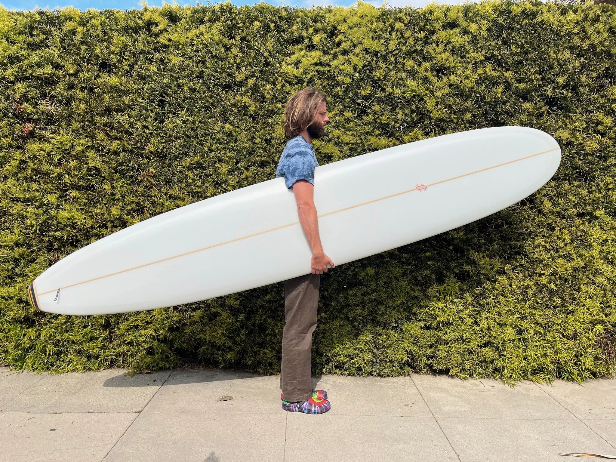 A man with shoulder-length hair and a beard is standing on a sidewalk holding a white surfboard with a signature near the center. He is wearing a blue patterned shirt, tan pants, and colorful tie-dye shoes. Behind him, there is a large green hedge.