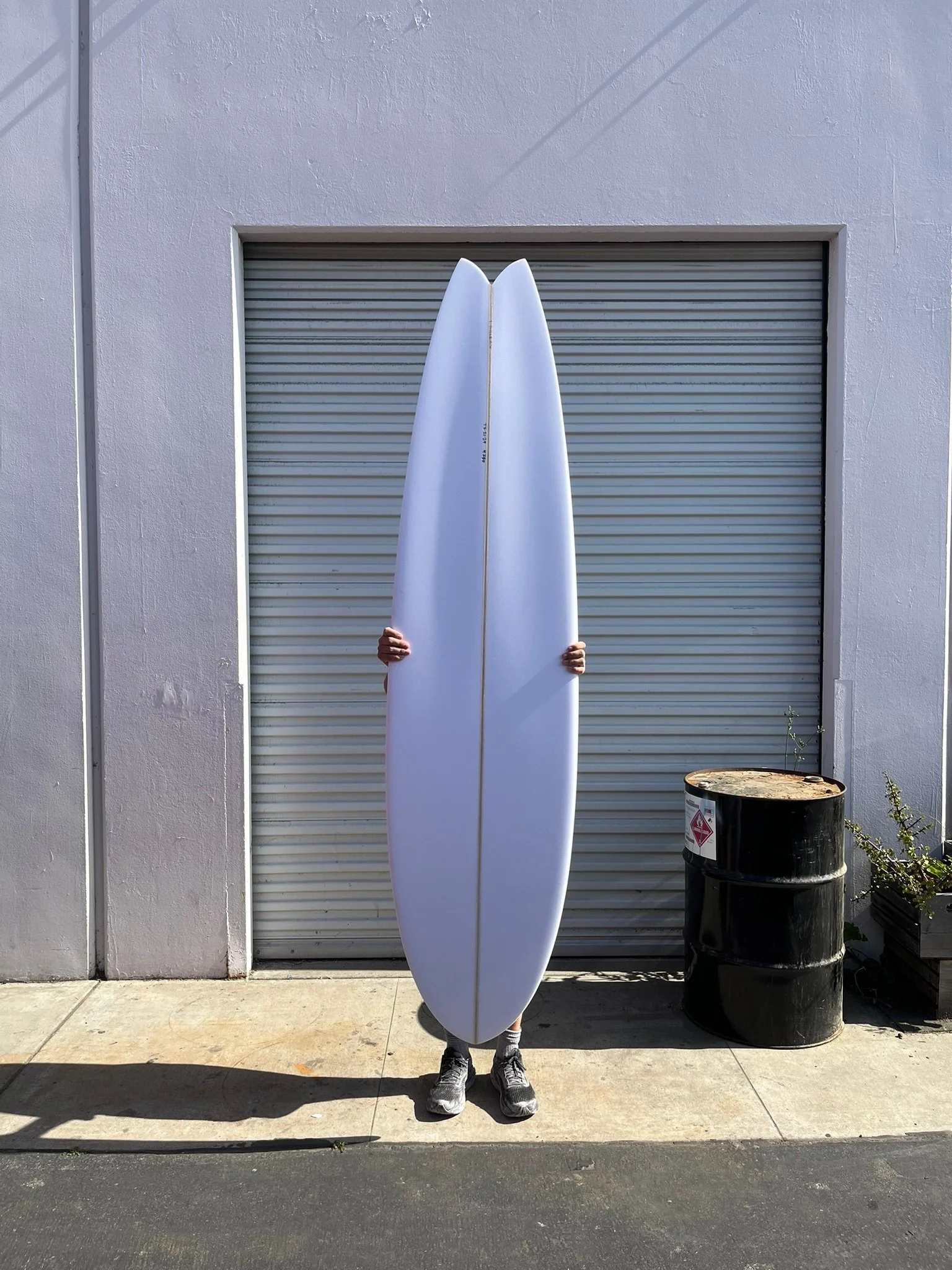 Person holding long fish style white surfboards in front of a building with a white wall and a grey roll-up door, near a black barrel with a warning label and some plants.