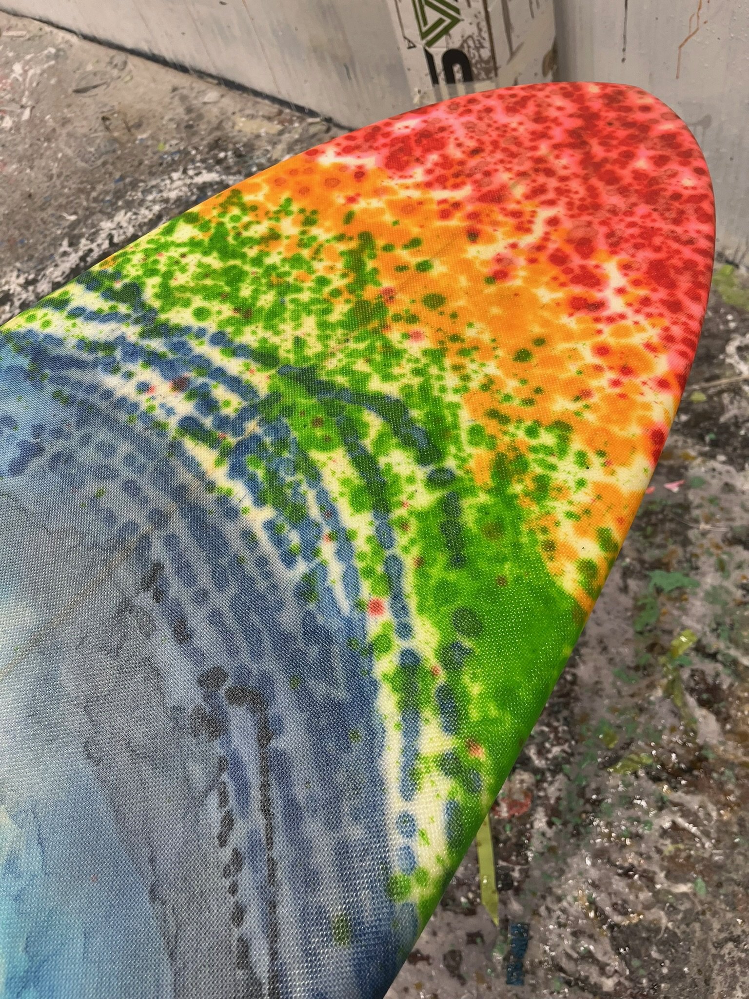Close-up view of a colorful, splatter-painted skateboard deck with red, orange, green, and blue paint, resting on the ground outdoors.