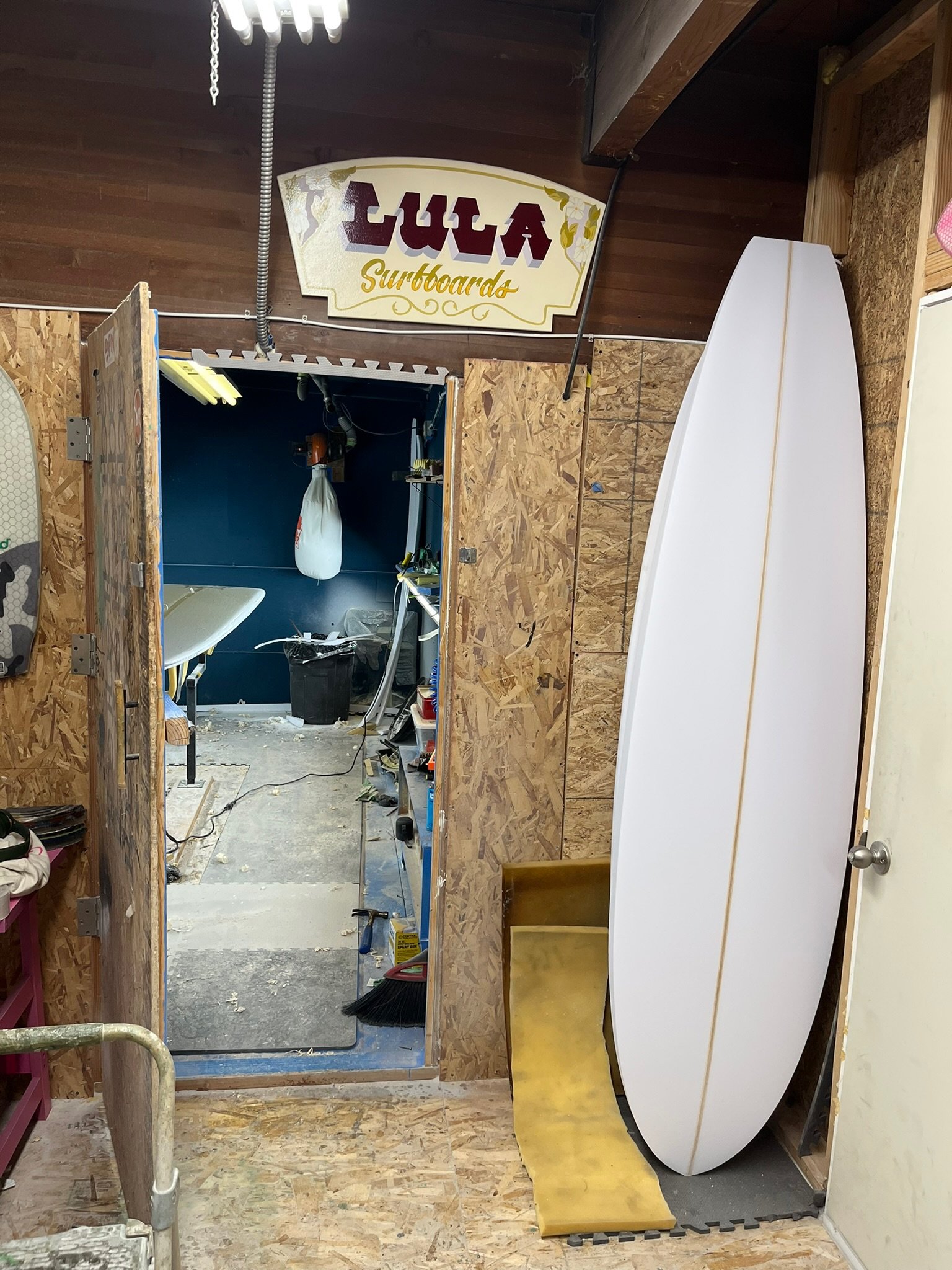 Interior view of a surfboard workshop with a large white surfboard standing upright on a yellow foam pad, a sign that reads 'LULA Surfboards' on the wall, and various tools and equipment used for surfboard shaping and construction.