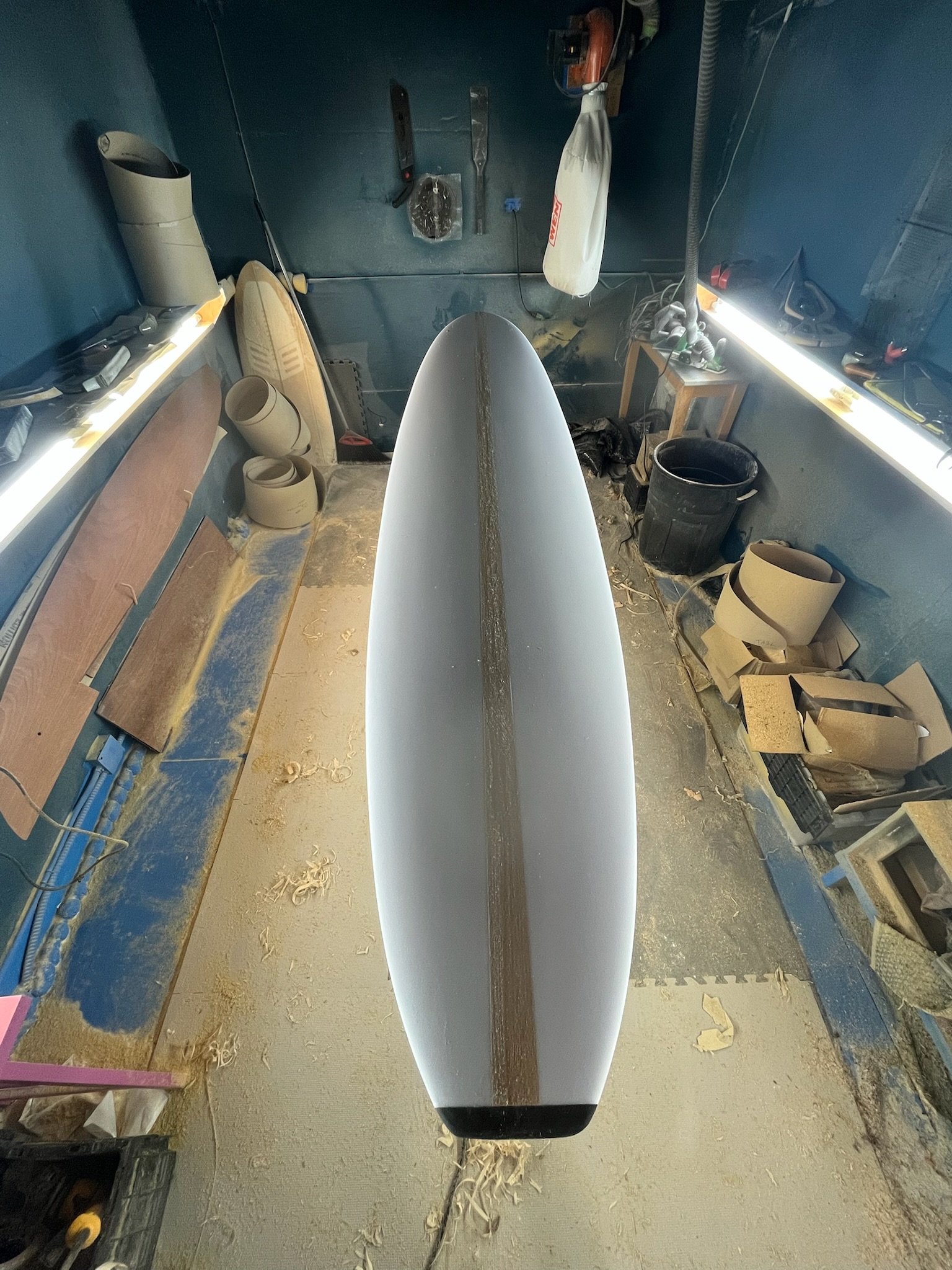 From a top-down perspective, the photo shows the nose of a surfboard inside a cluttered workshop. The workshop contains tools like files and a hammer, a workbench with a strip of bright light, cardboard tubes, and a trash bin. The floor and walls sho