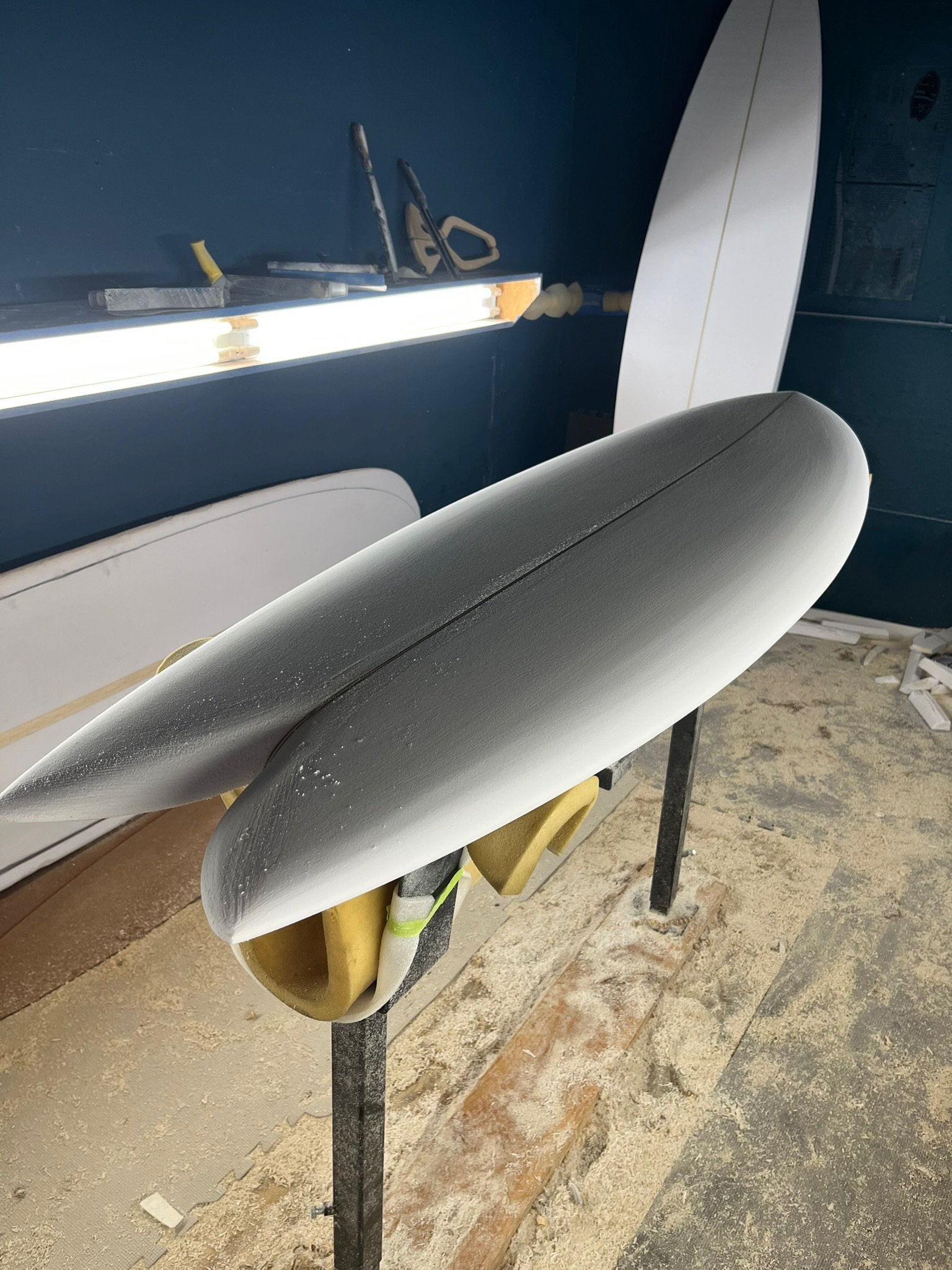 Fish surfboard design in a shaping bay with tools on a dark walls and surfboard foam on the floor.
