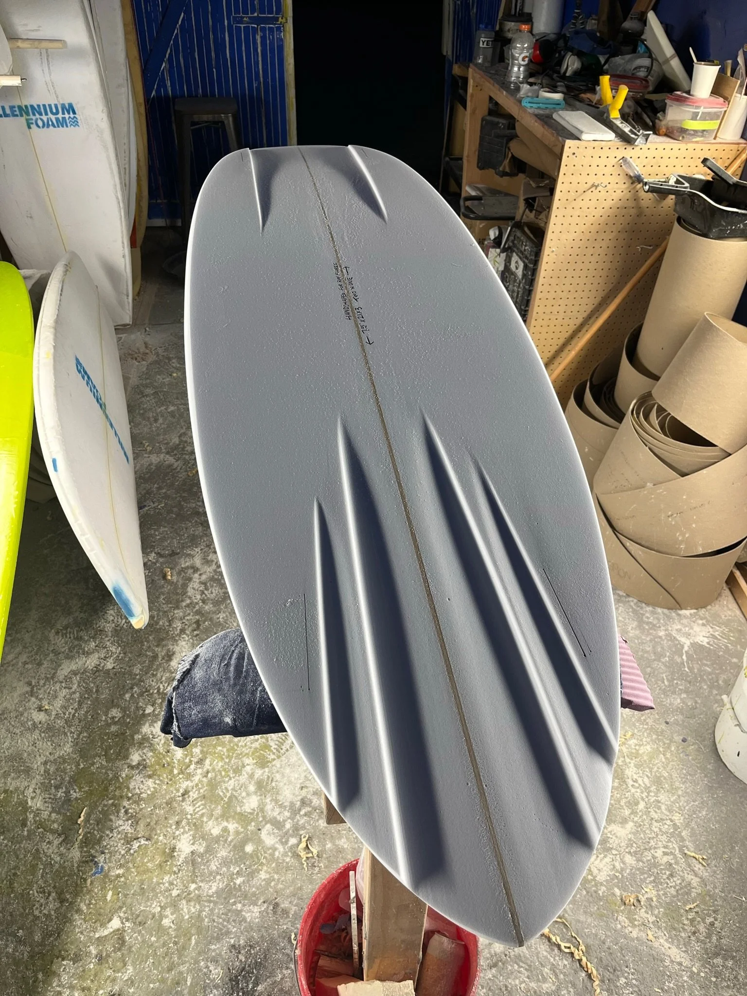 A surfboard with a metallic finish mounted on a wooden stand in a workshop, surrounded by various tools, supplies, and rolls of paper. Displaying channel bottoms design.