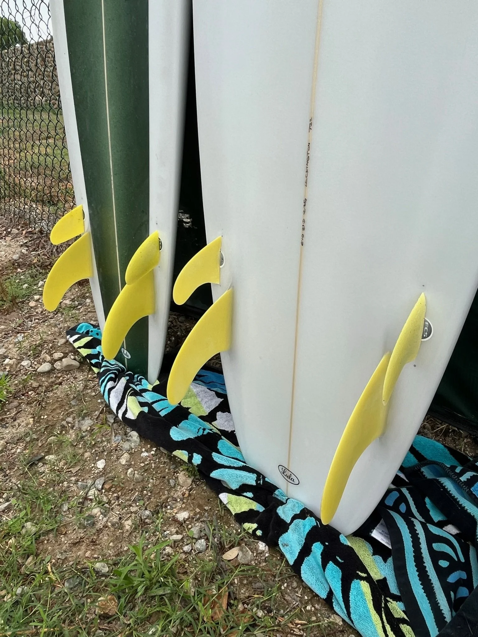 A row of surfboards with colorful yellow fins and various designs, placed vertically on a striped towel on the ground outdoors. There is a chain-link fence and some rocks and grass in the background.